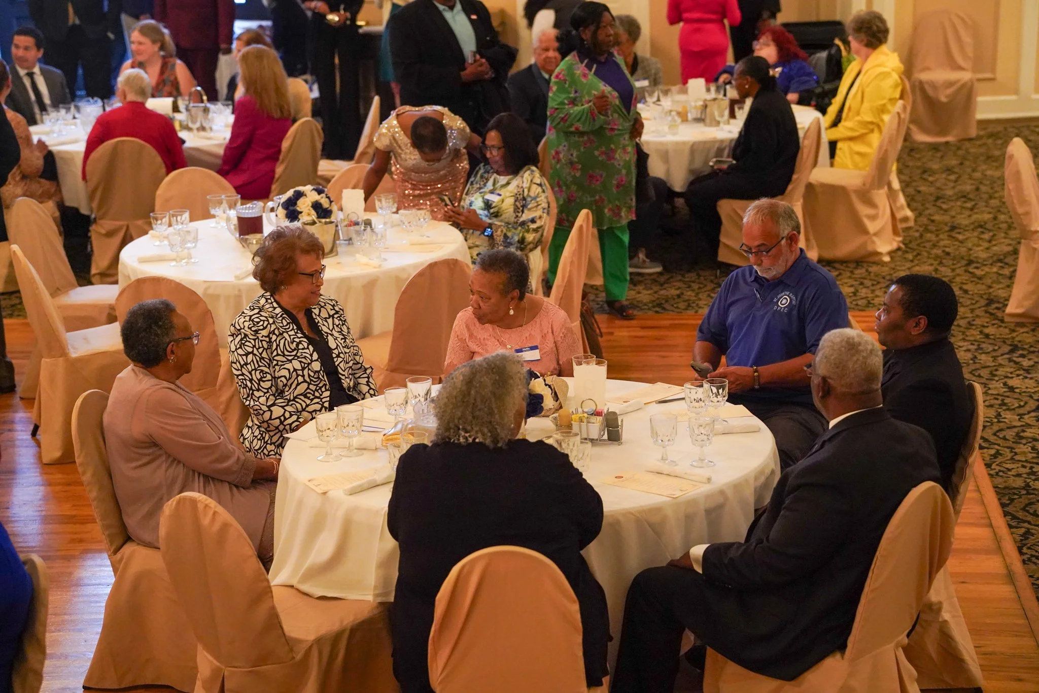 People gathered around decorated tables at a formal event in a banquet hall, engaging in conversations and using mobile devices.