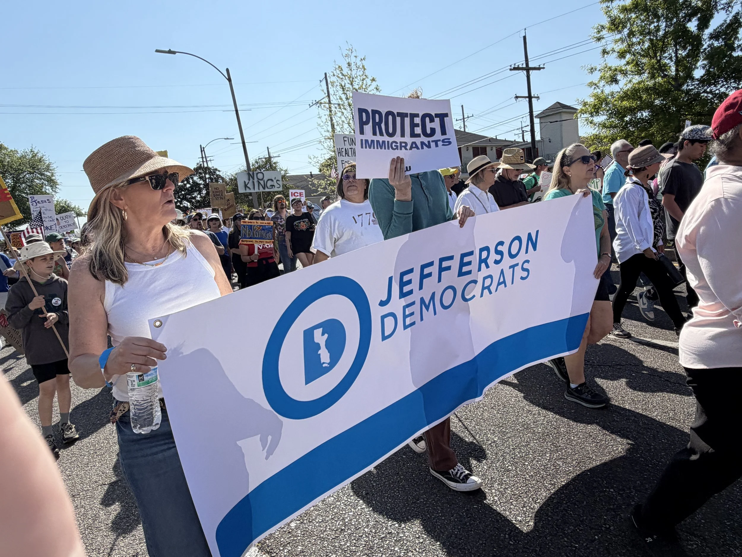 A diverse group of people participating in a protest march on a sunny day, holding signs with messages like "Protect Immigrants" and other slogans advocating for immigrant rights. The crowd includes women wearing hats and sunglasses, and a large banner with a blue logo and text.