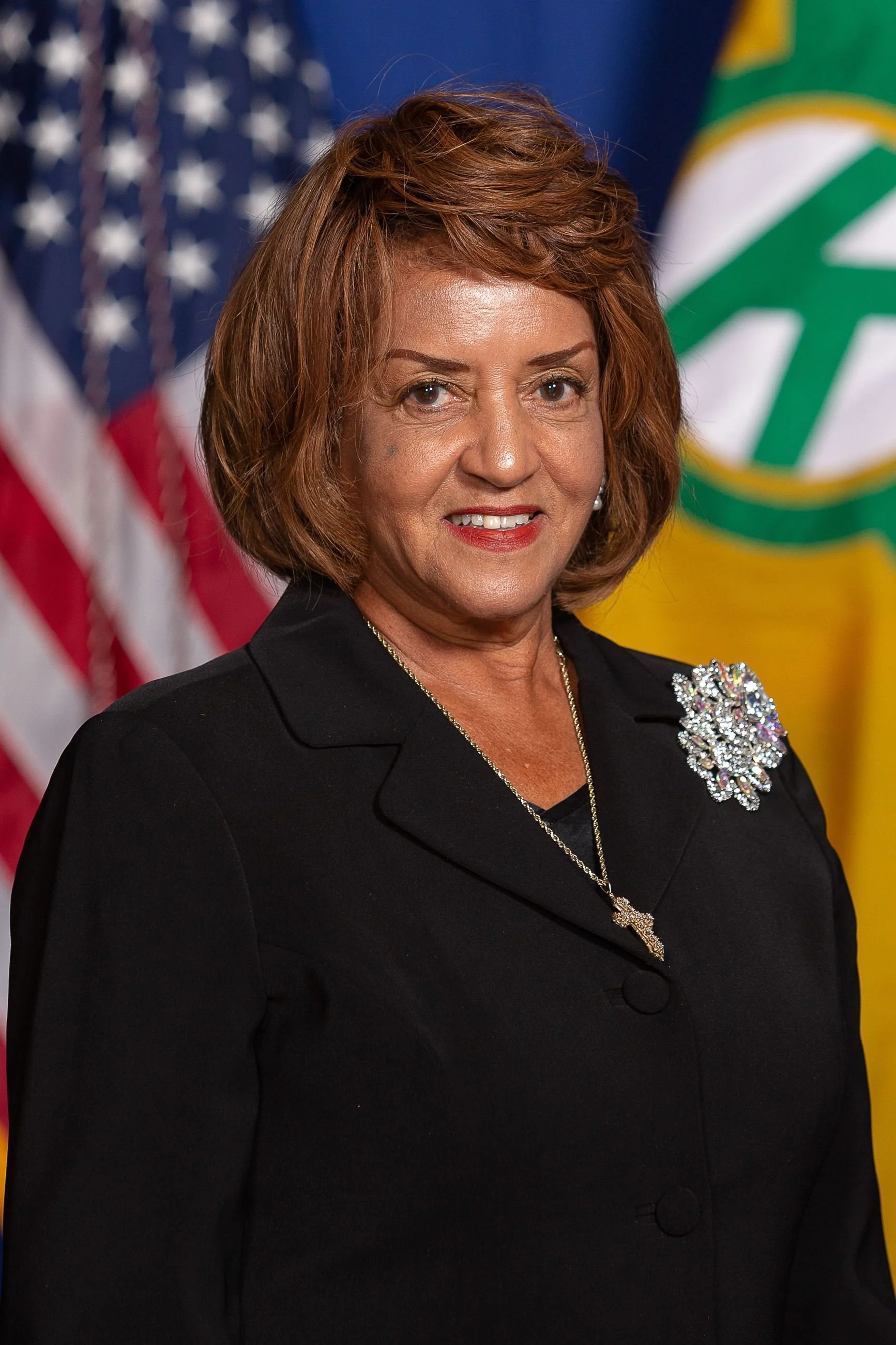A woman with short reddish-brown hair wearing a black blazer with a large, sparkly brooch on the shoulder, standing in front of American and other flags.