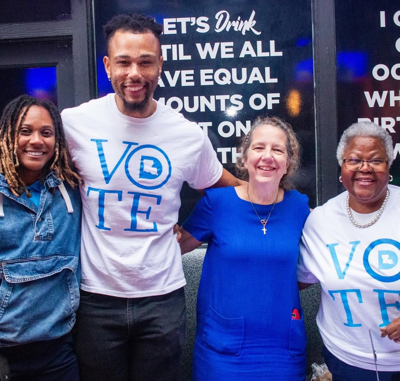 Group of four smiling women and one smiling man standing together at an event, with a dark background and partially visible sign with white text.