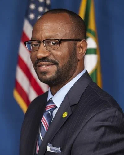 A man wearing glasses, a dark suit, and a striped tie, standing in front of American and another flag, during a formal event or photo shoot.