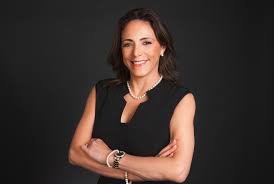 A woman with shoulder-length dark hair smiling, wearing a sleeveless black top and a pearl necklace, standing against a gray background.