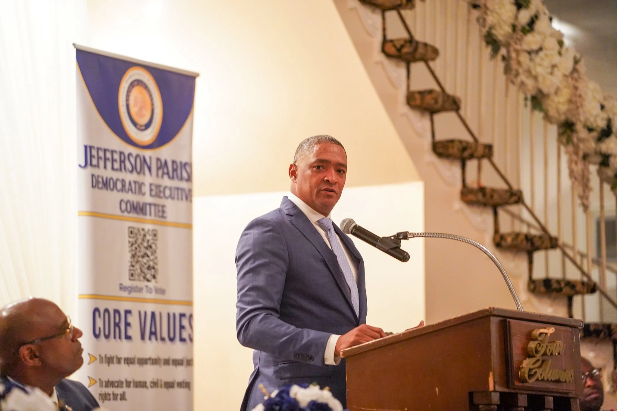 A man in a blue suit and tie is speaking at a podium at an event. Behind him is a banner that reads 'Jefferson Parish Democratic Executive Committee' with a QR code and the slogan 'Core Values.' The setting appears to be a formal indoor venue with floral decorations and a decorative balcony.