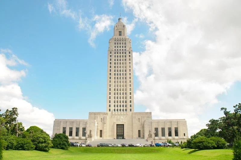The Texas State Capitol building with a green lawn and trees in front, under a partly cloudy sky.