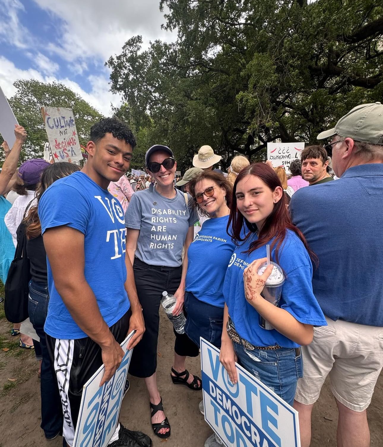 Group of four smiling people participating in a protest or rally, holding signs and wearing blue shirts with messages about democracy and human rights, outdoors with trees and other protesters in the background.