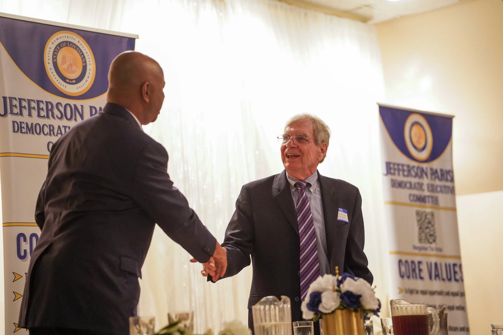 Two men shaking hands at an event, with banners displaying 'Jefferson Parish Democratic Committee' and 'Core Values' in the background. One man is bald and wearing a dark suit, the other has glasses and gray hair, wearing a dark suit and striped tie.