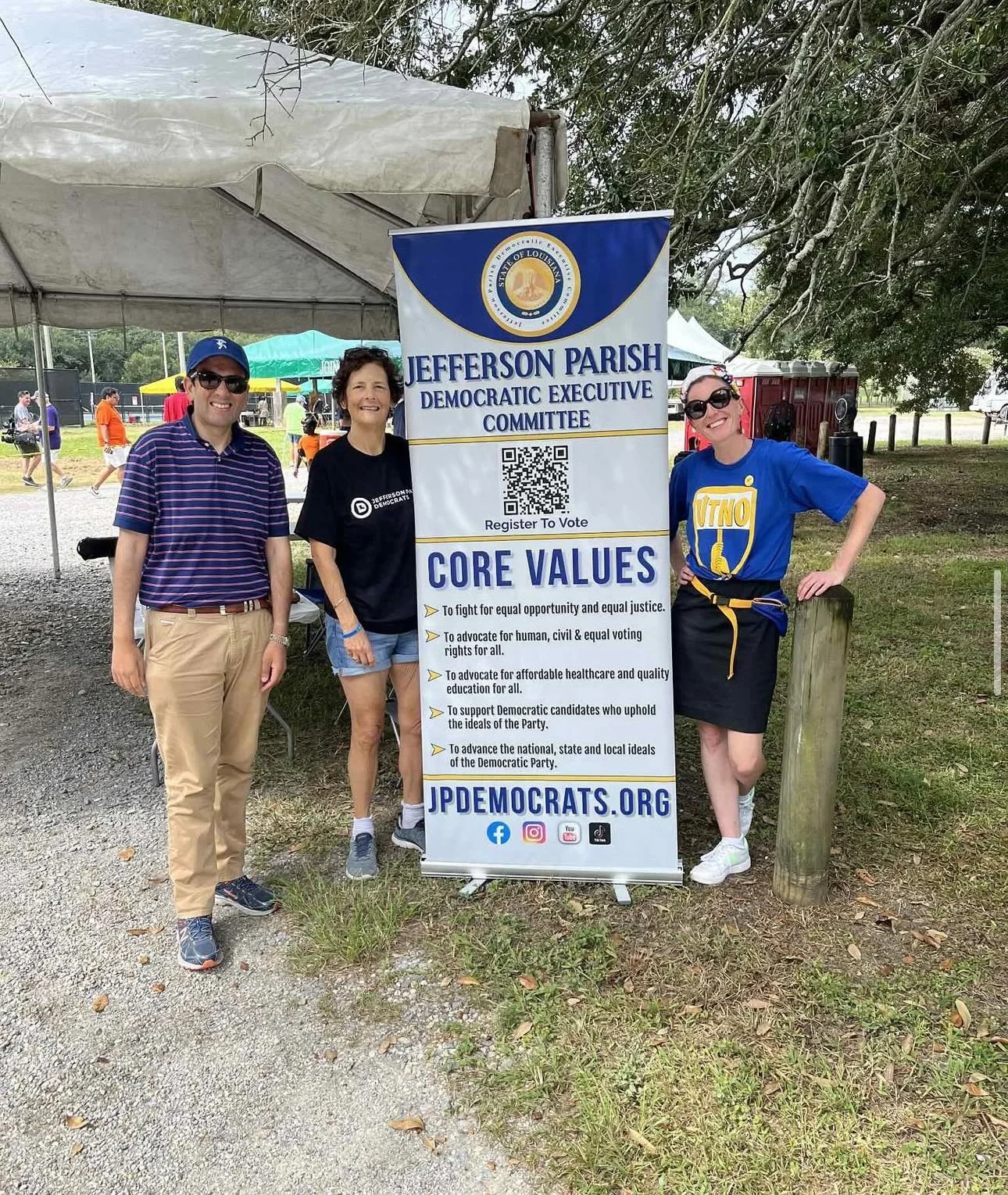 Three people standing next to a banner for Jefferson Parish Democratic Executive Committee at an outdoor event. The banner displays core values advocating for equality, voting rights, healthcare, and supporting Democratic candidates.