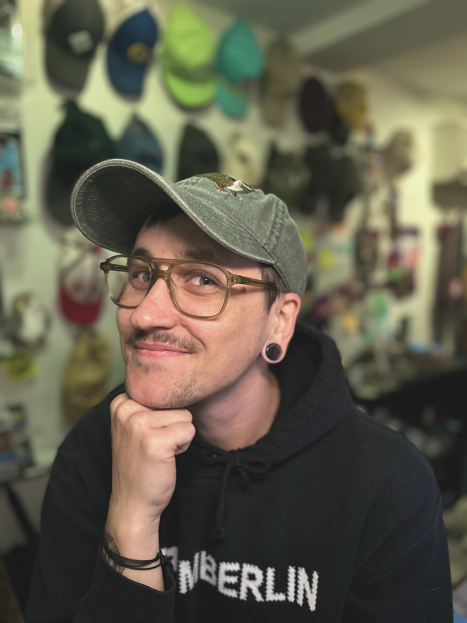 Image of Clair wearing a baseball cap and glasses, smiling with chin resting on hand.