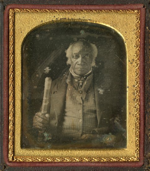 An elderly Black man sits for a portrait, dressed in nineteenth century clothing and holding a cane. His portrait is enclosed in a gold frame.