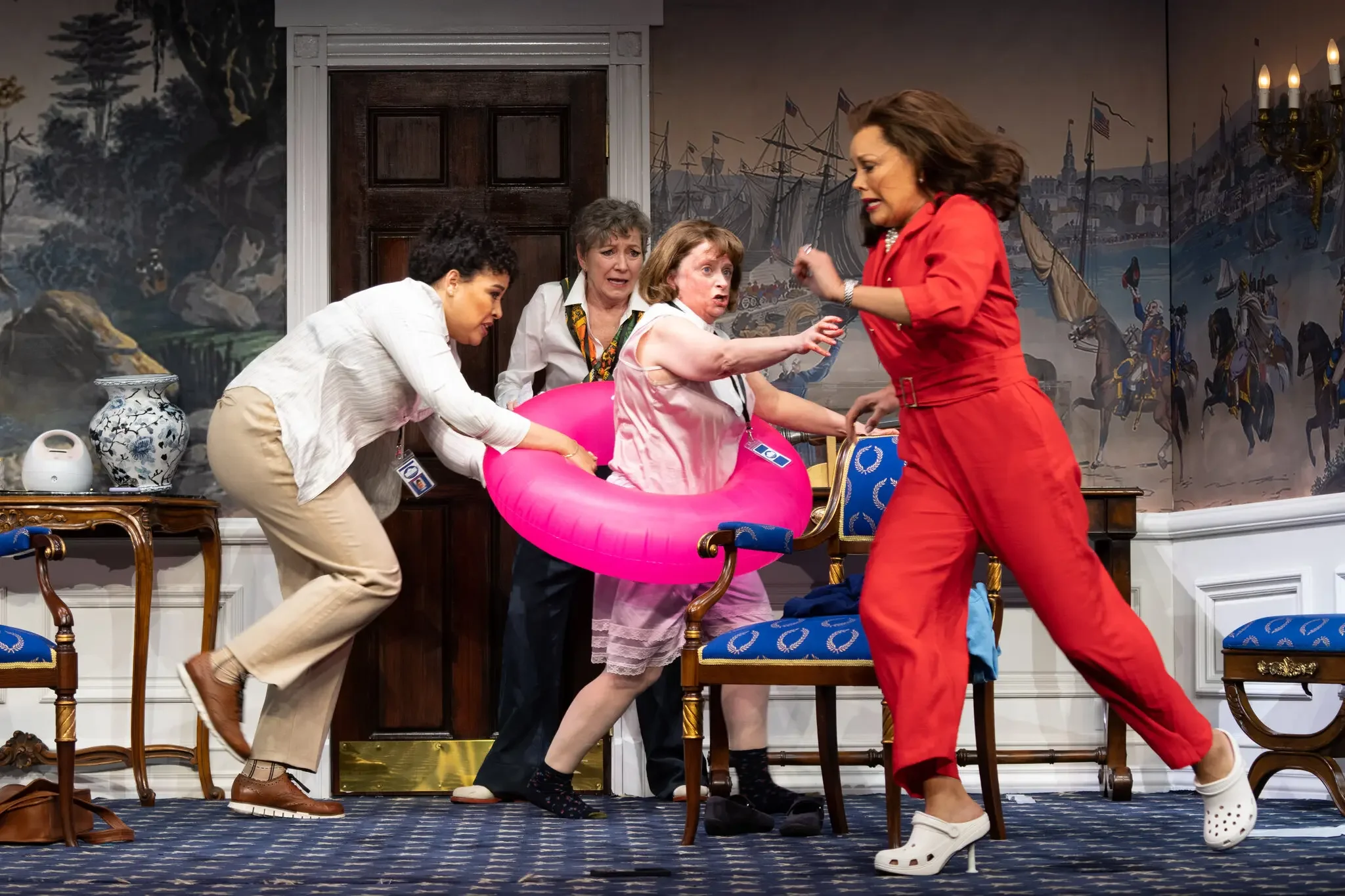 In the center of the photo is a woman in a slip with a pink inner tube around her, another woman is holding the inner tube, while one behind is behind her and one in a red pantsuit and high heeled crocs runs in front of them all