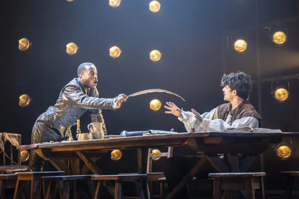 a black man with a quill leangs over a table talking to another man. both are in elizabethan costume.