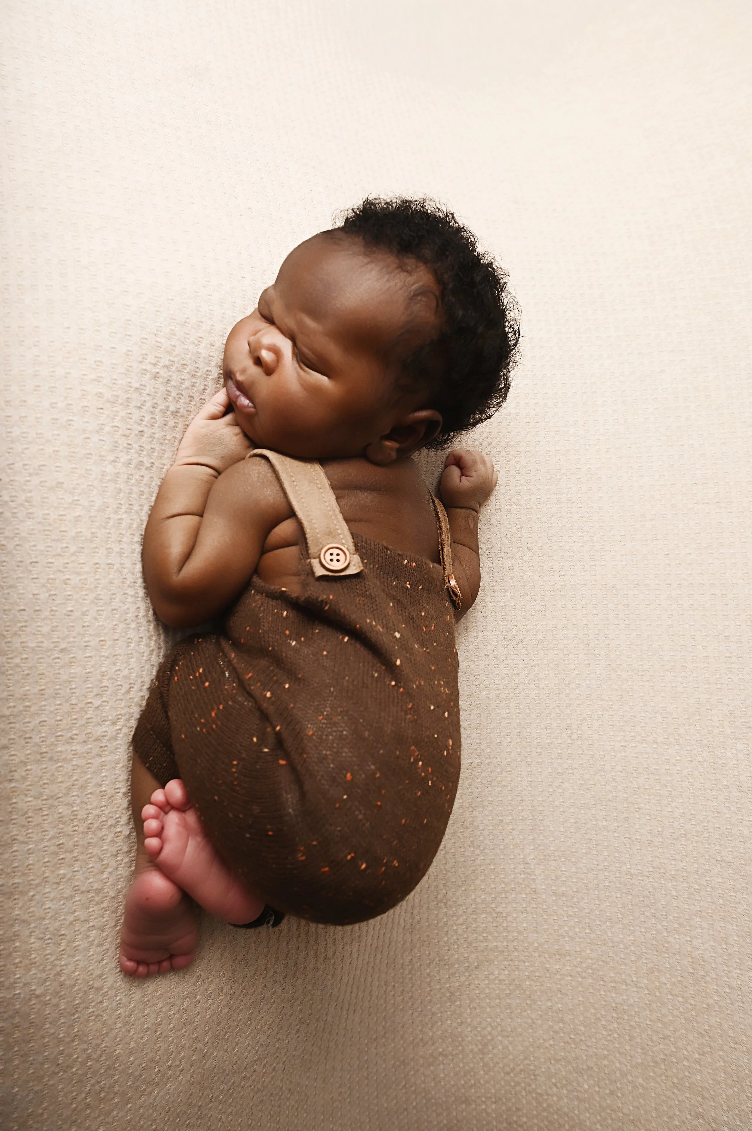 Overhead view of a newborn baby sleeping in a brown romper on a neutral textured backdrop in a Shreveport studio.