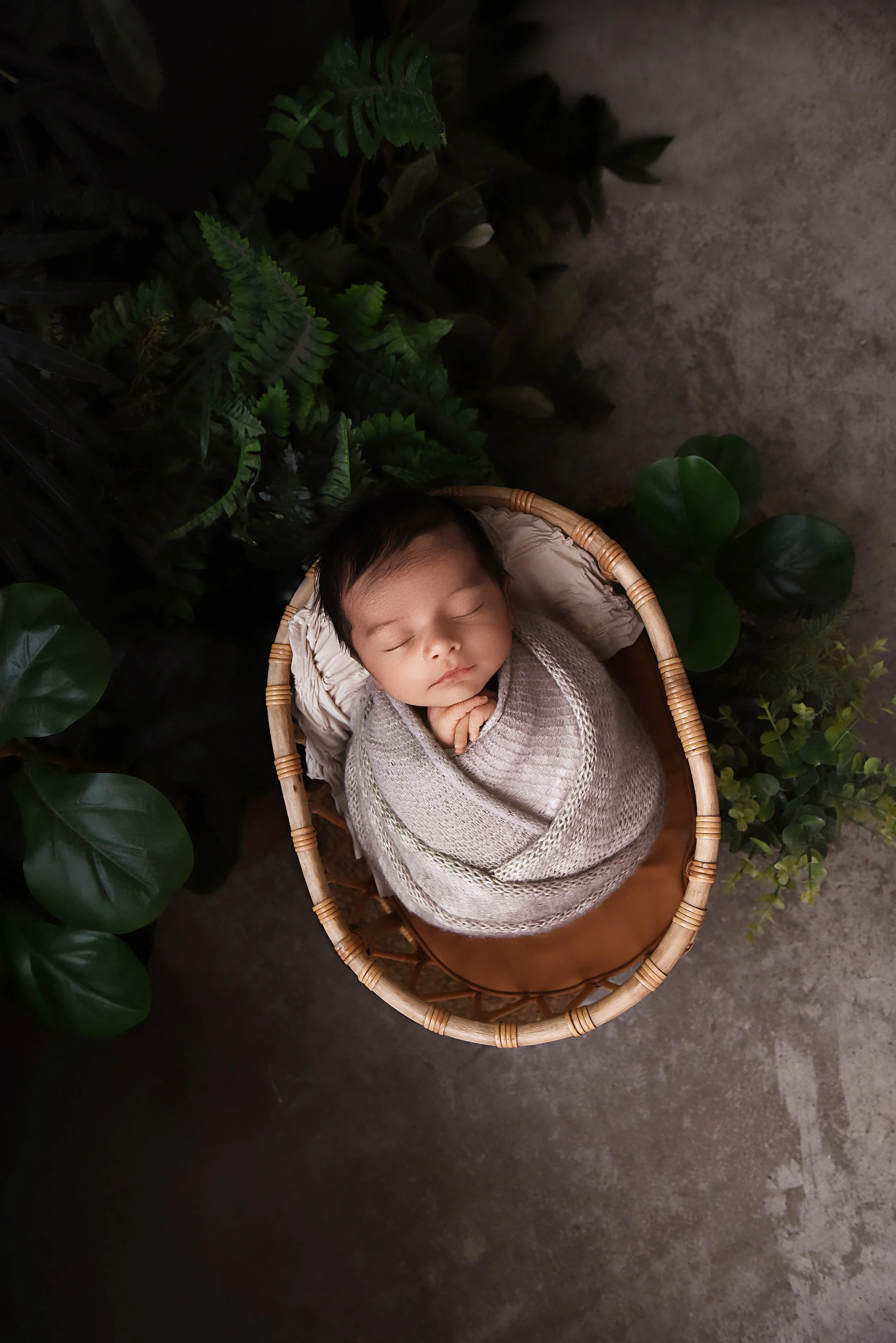 newborn wrapped in taupe knit in a basket surrounded by greenery