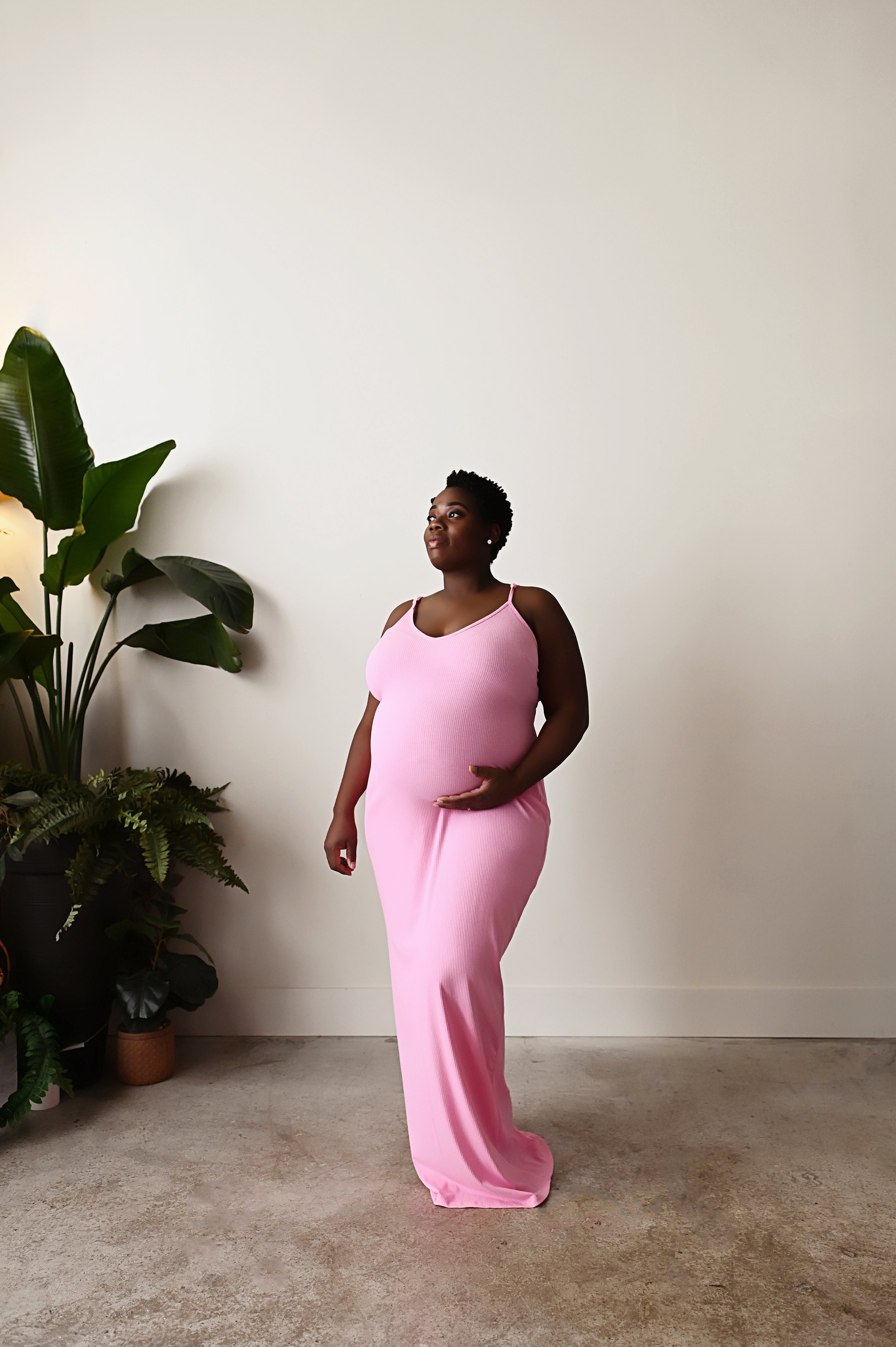 pregnant mother wearing pink maternity dress standing near plants during studio maternity photography session