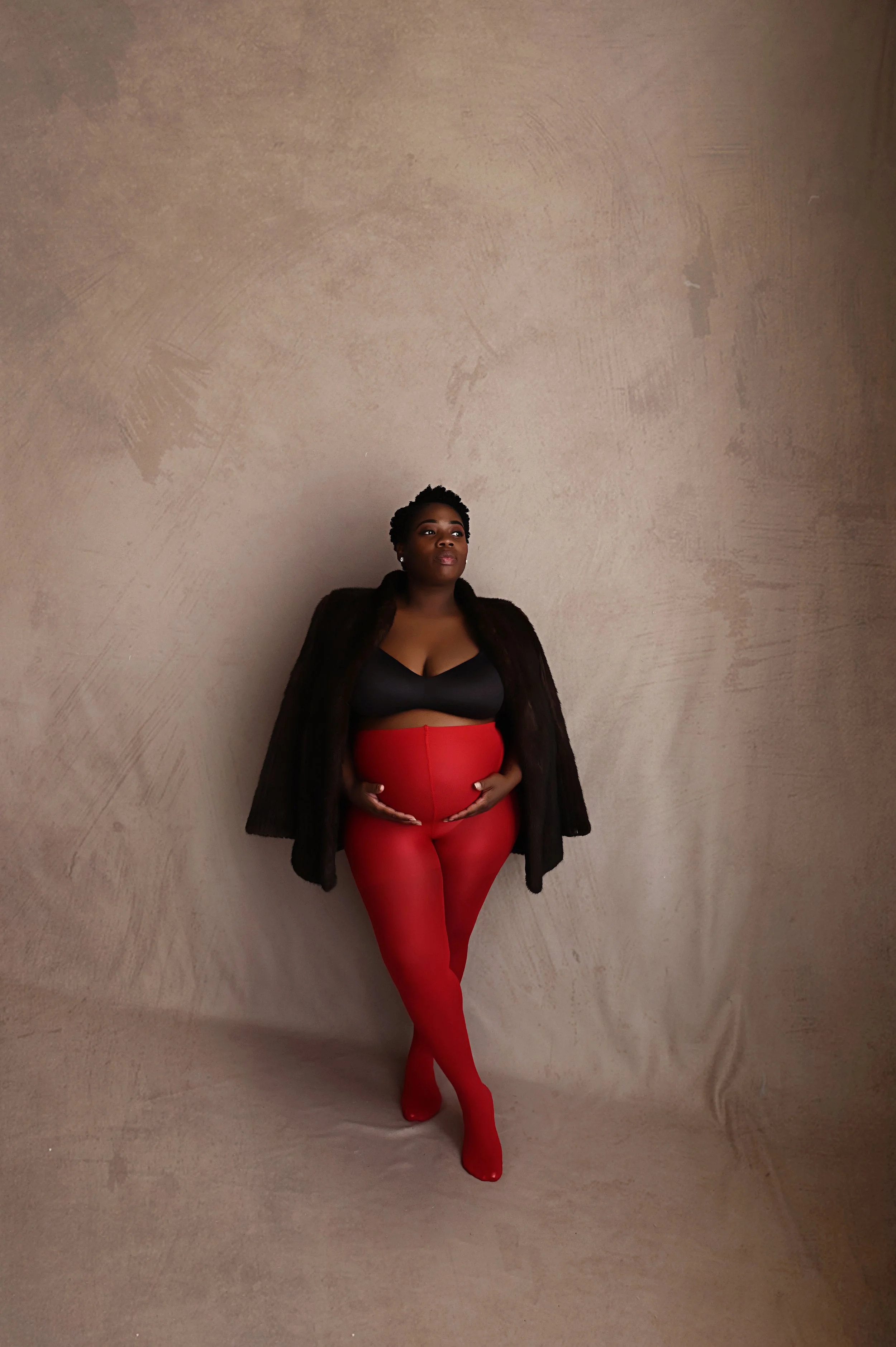 pregnant mother wearing red tights and black wrap posing against neutral backdrop during modern maternity photography session