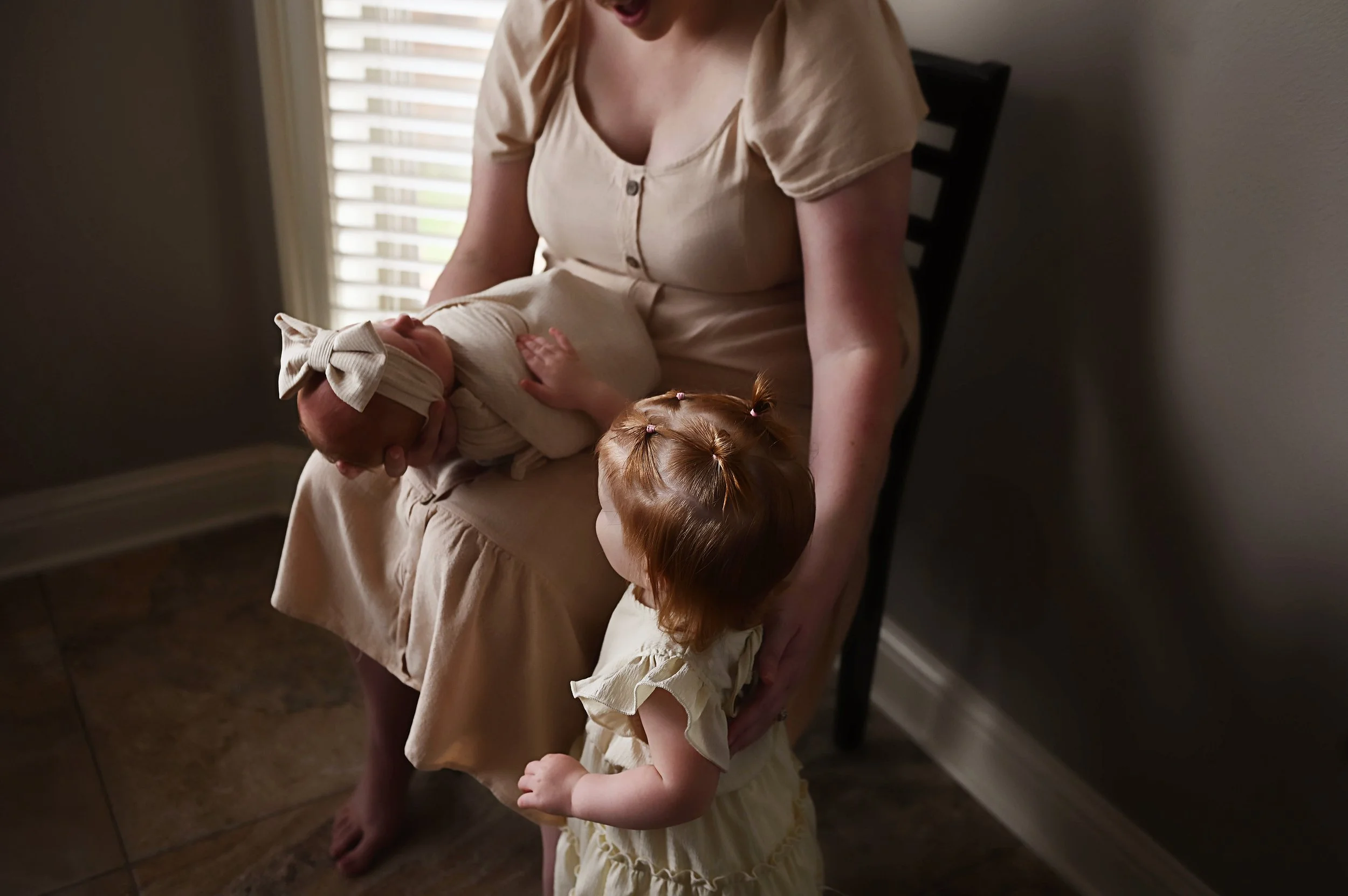 Toddler reaching out to touch newborn sister held in mother’s lap during an in-home newborn photography session.