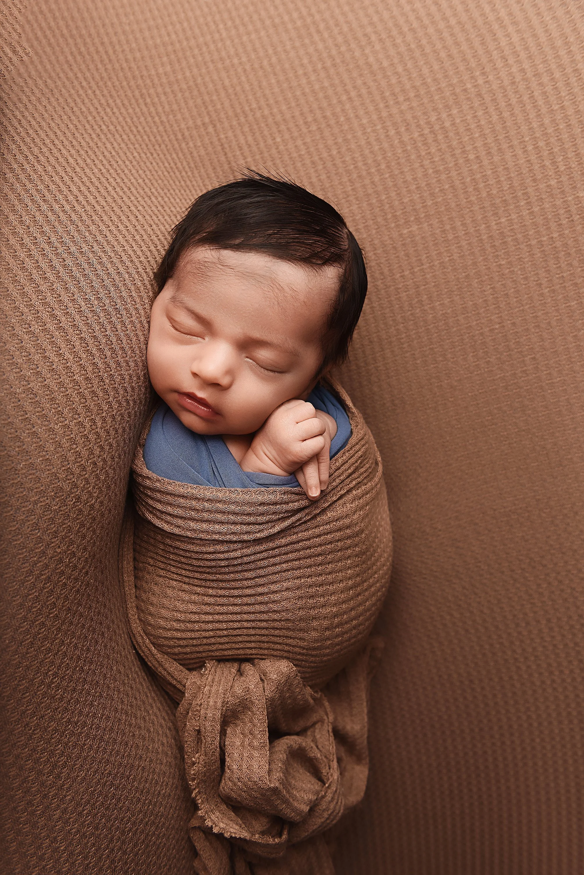 sleeping newborn wrapped in brown fabric with a blue under layer on textured backdrop