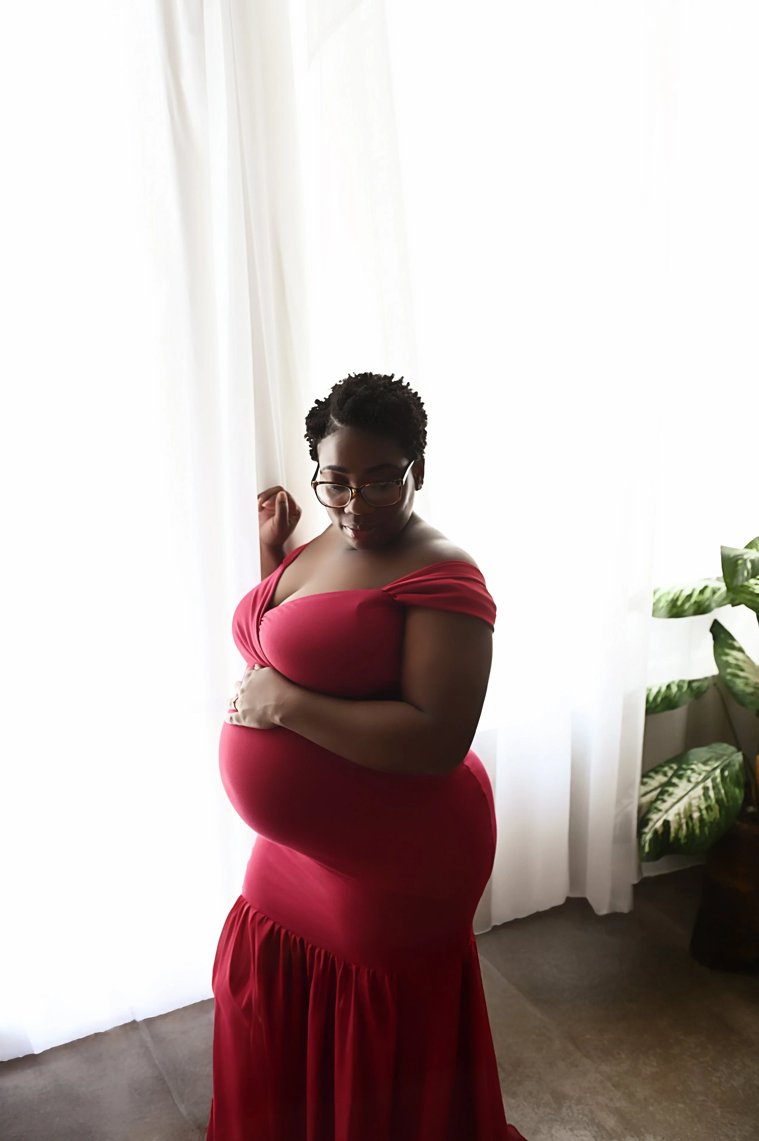 pregnant mother wearing a red maternity dress standing beside sheer window light during a maternity photography session in Shreveport Louisiana