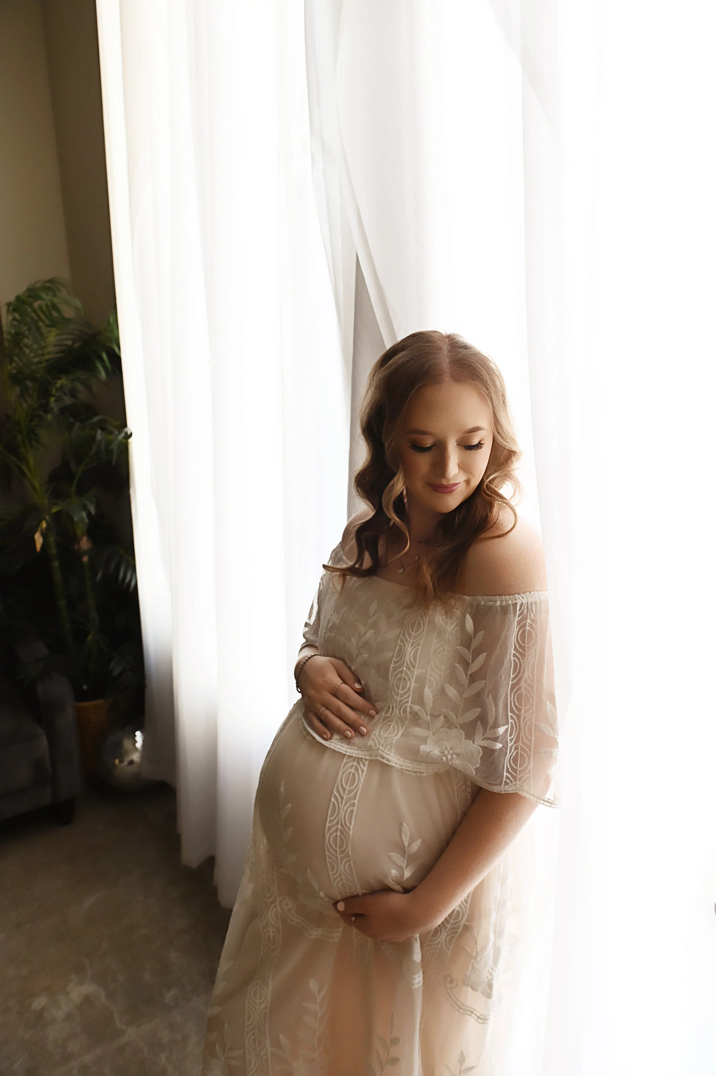 Studio maternity photo of an expecting mother in a cream lace gown standing by a window with natural light