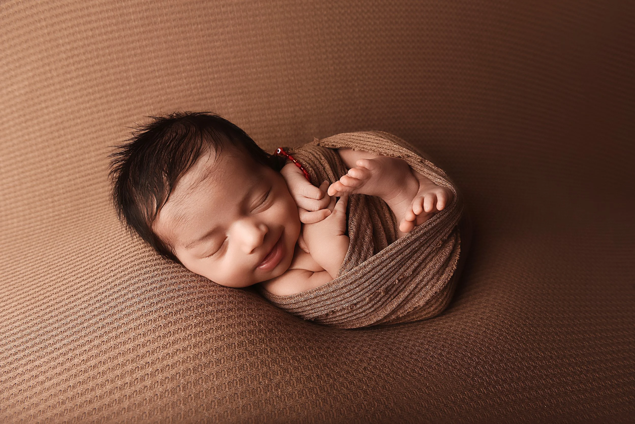 newborn baby wrapped in brown fabric smiling softly while curled up during studio session
