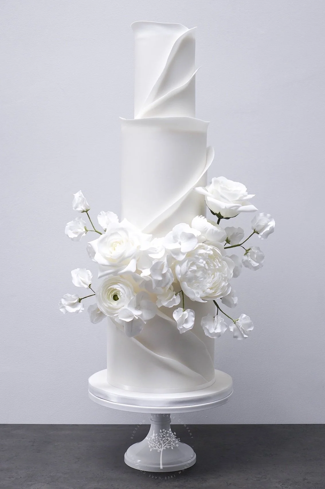 Modern three‑tier white wedding cake with soft spiral ruffles and a romantic cluster of handcrafted sugar roses, peonies, created by Blossom Tree Cake Company in Harrogate for luxury Yorkshire weddings at venues such as Grantley Hall.