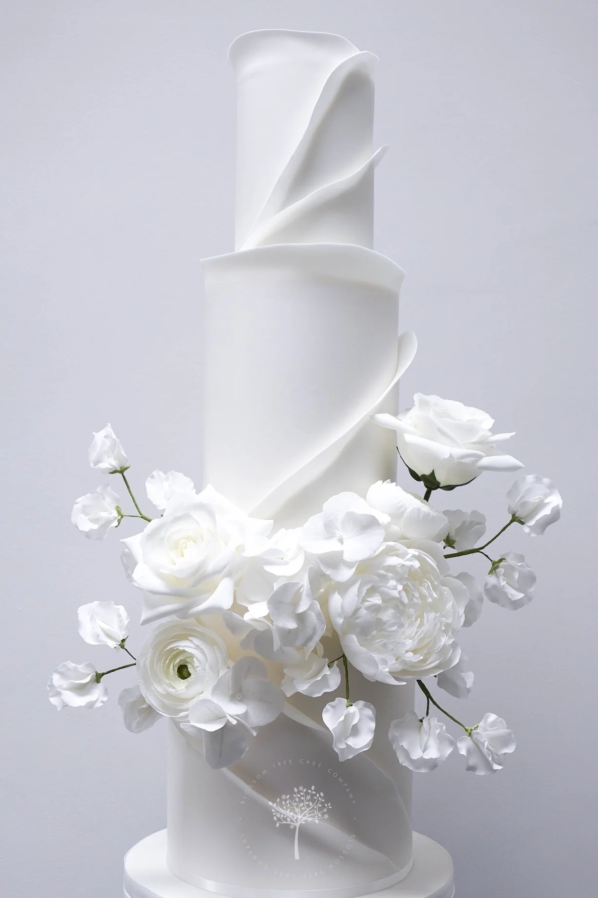 Modern multi‑tier white wedding cake with sculpted spiral detailing and a romantic cluster of handcrafted sugar roses, peonies, and hydrangeas, created by Blossom Tree Cake Company in Harrogate for luxury Yorkshire weddings at Grantley Hall.