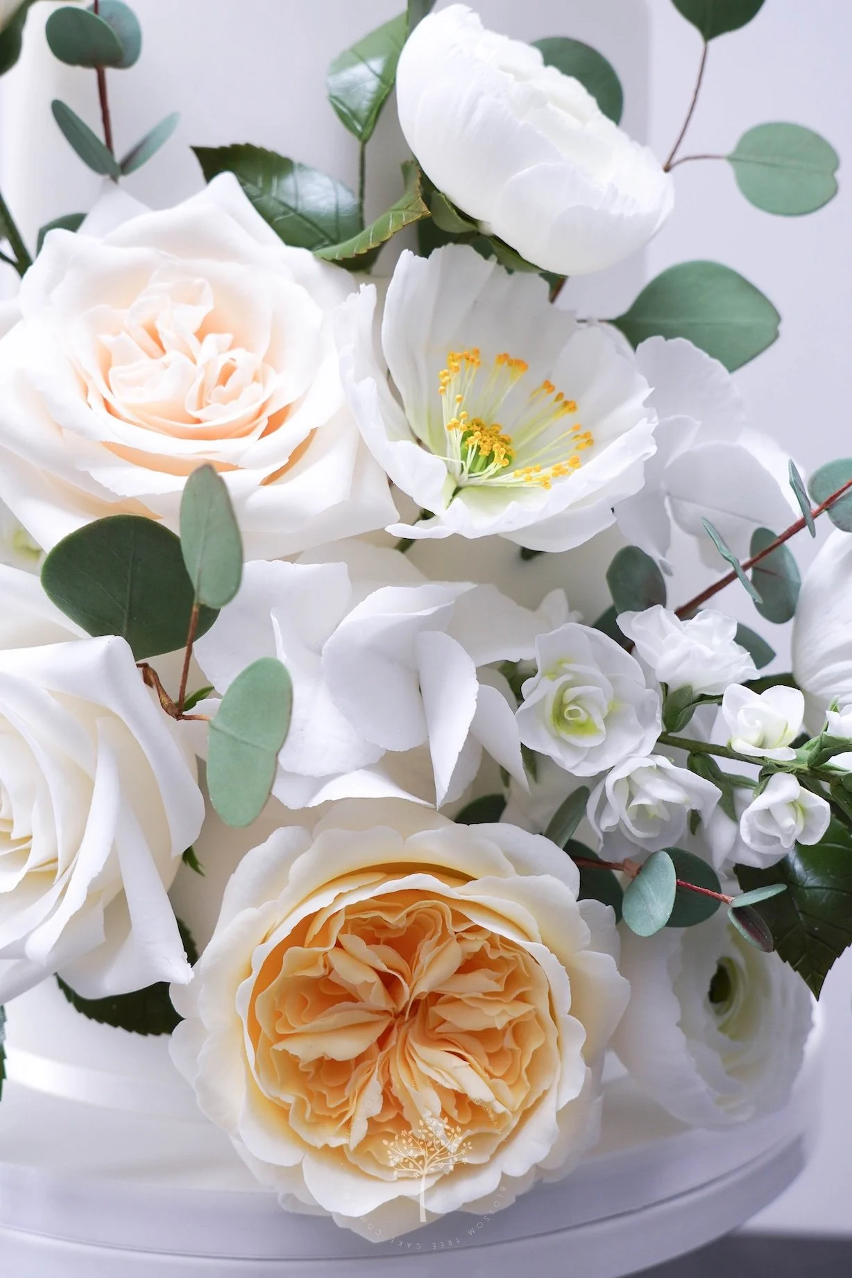 Close‑up of an elegant Blossom Tree Cake Company wedding cake decorated with soft, romantic sugar flowers, including white and pale peach roses, white peonies, delicate blossoms, and eucalyptus foliage, showcasing refined handcrafted floral detail.