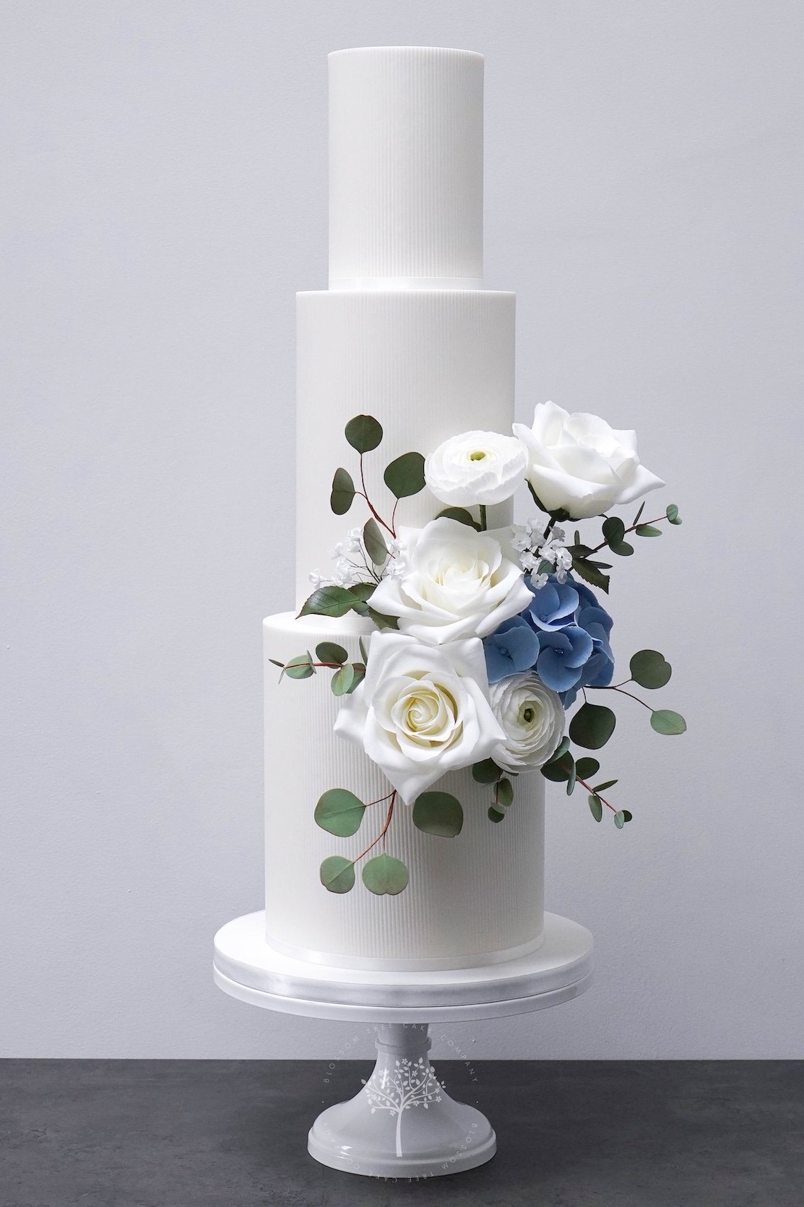 Three-tier white wedding cake with soft textured tiers with handcrafted sugar roses, ranunculus, blue hydrangeas and eucalyptus, created by Blossom Tree Cake Company in Harrogate for luxury Yorkshire weddings at Goldsborough Hall.