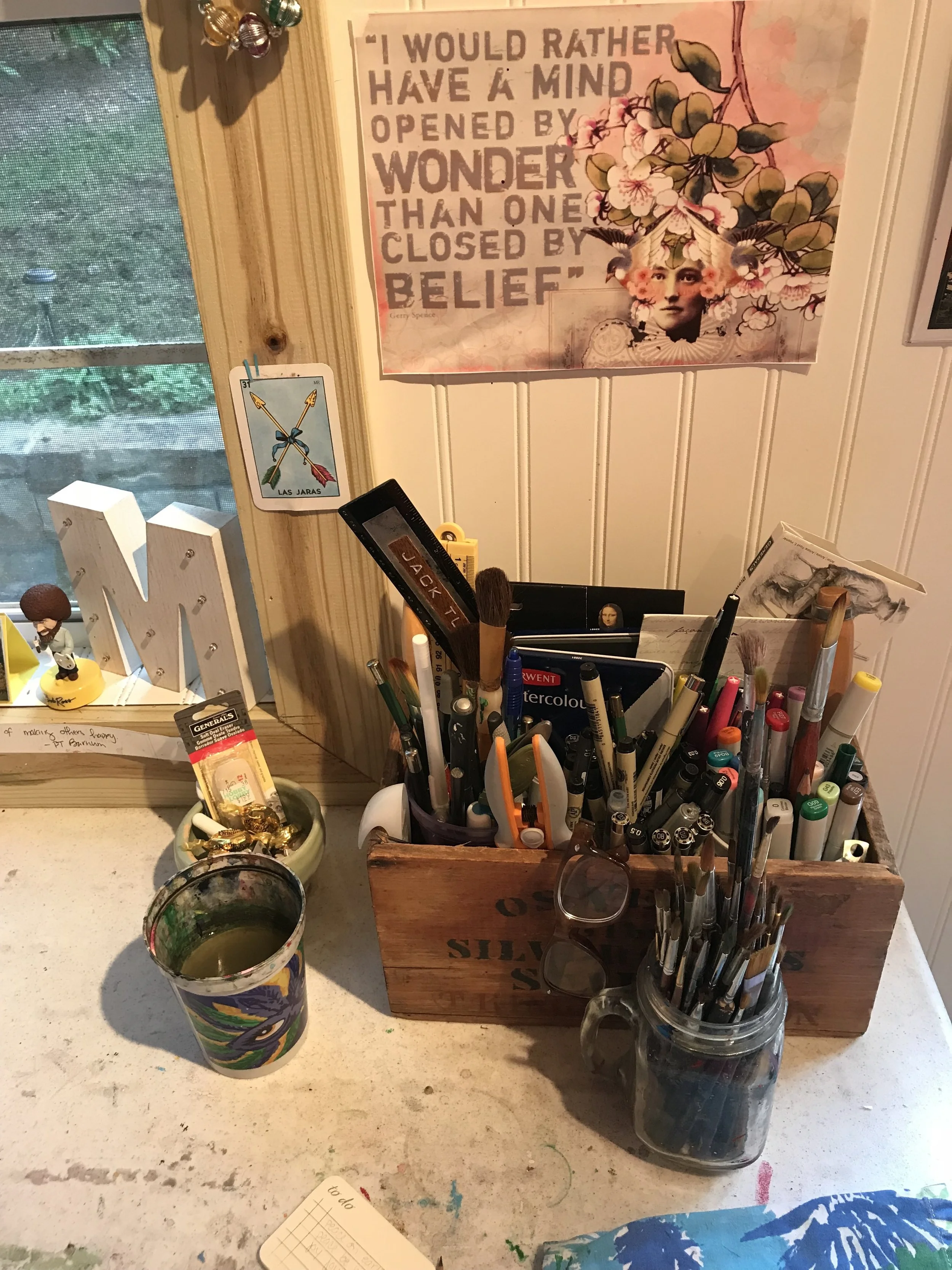 This little corner of my desk holds all of my essentials - brushes, water, black ink pens, copic markers, scissors, rulers and erasers. The little green bowl was my only successful attempt at making something on a pottery wheel. It holds my glasses, erasers, pencil sharpeners and werther’s originals. My Bob Ross bobblehead is one of my favorite toys. He has an easel where I can switch out his paintings in miniature. When I’m feeling sassy, I turn on my marquee M. The quote above my supply box is one of my favorites.