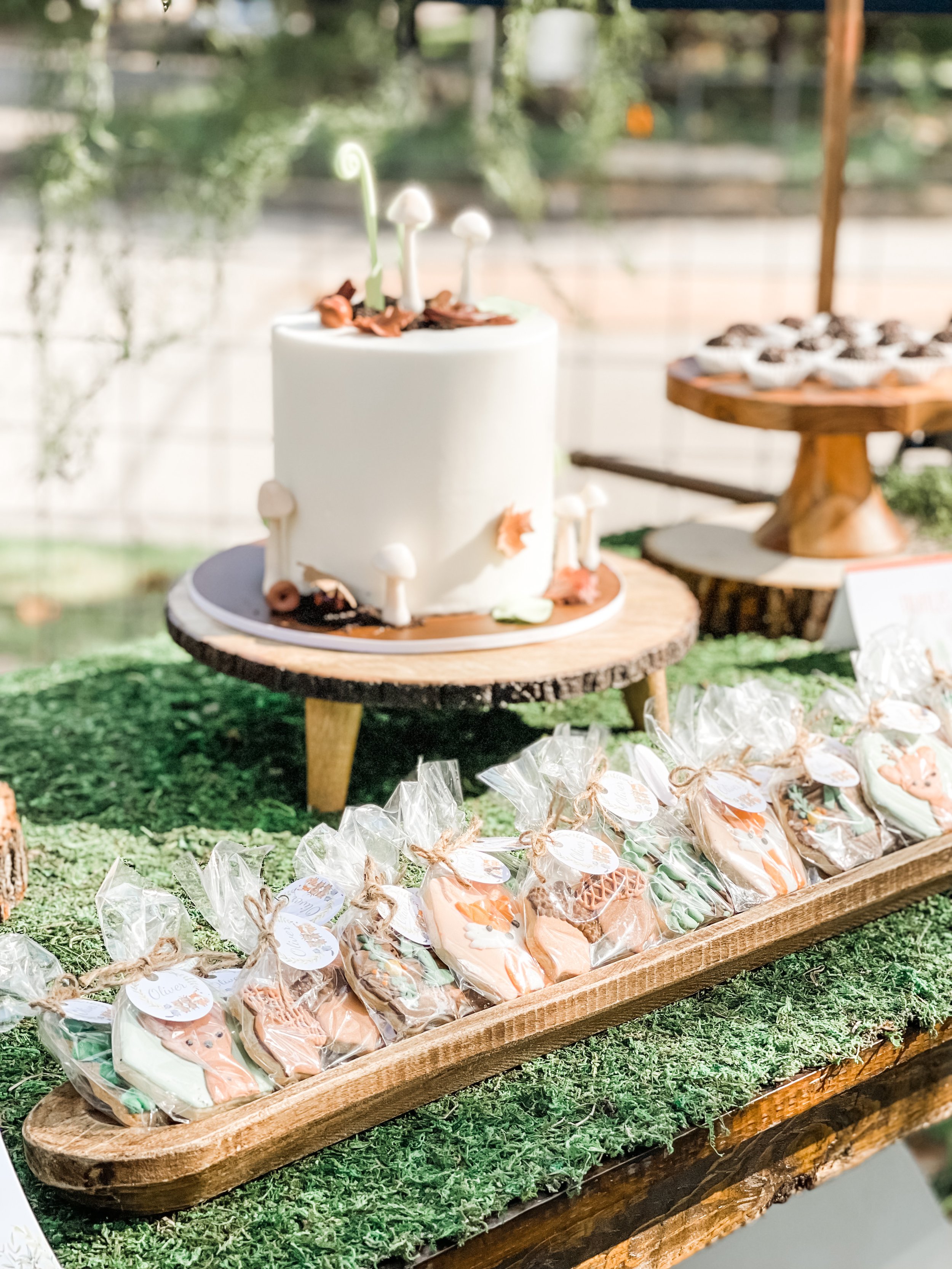  Woodland-themed first birthday dessert table with white frosted cake and fondant mushroom, plant, and leaf decorations on a wood slice pedestal stand, with woodland-themed iced cookie favors in individual plastic bags 
