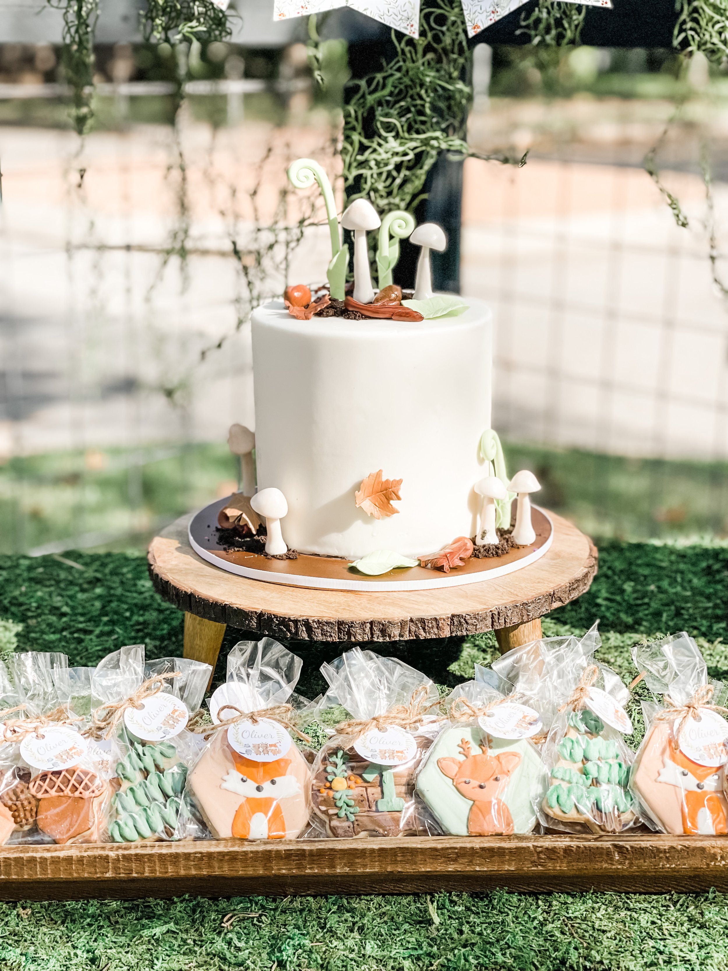  Woodland-themed first birthday cake with white frosting and fondant mushroom, plant, and leaf decorations on a wood slice pedestal stand, with woodland-themed iced cookie favors in individual plastic bags 