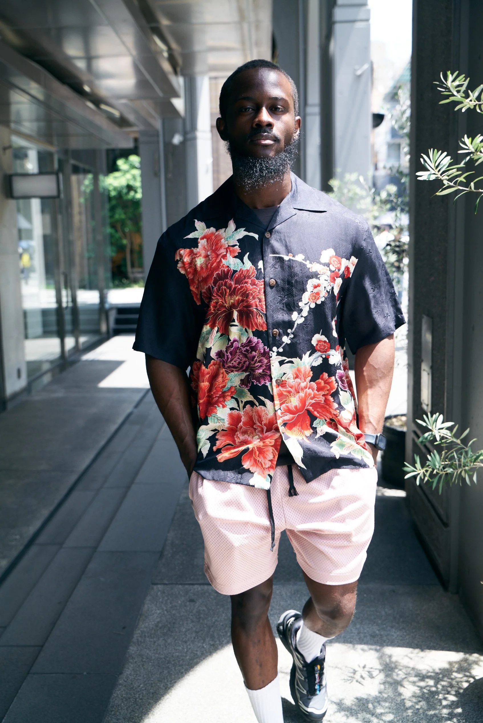 A man walking outdoors wearing a colorful Fukika "Royal Peony" silk aloha shirt, light pink shorts, sneakers, and a watch, with hands in his pockets, in a modern urban setting with sunshine.