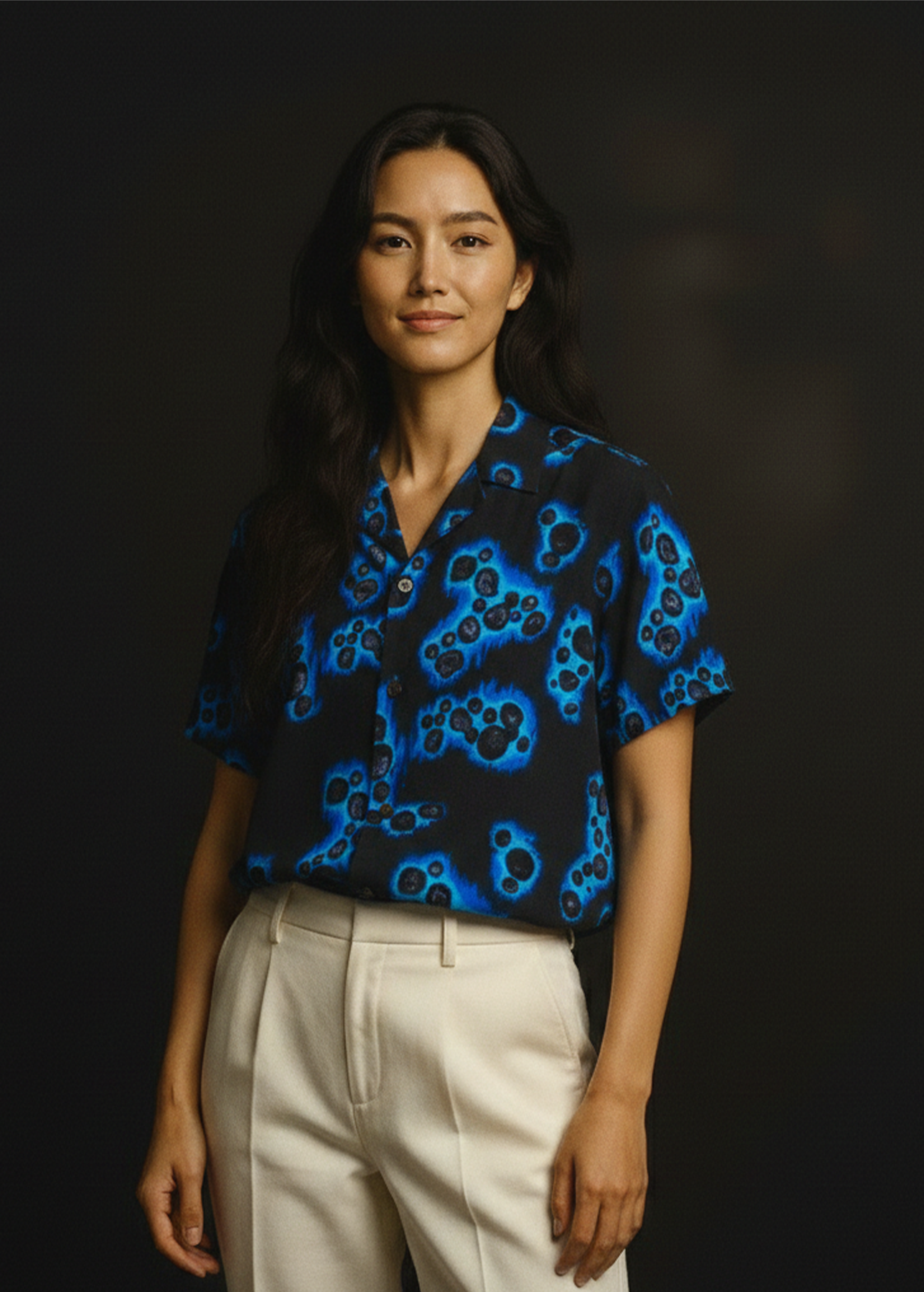 A woman with long black hair wearing a "The National Treasure Yohen Tenmoku patterned silk aloha shirt and beige pants, standing against a dark background.