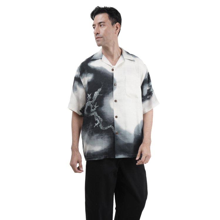 A young man wearing a " Dragon flying over moung Fuji" silk aloha shirt with a black and white abstract design and black pants, standing against a plain white background.