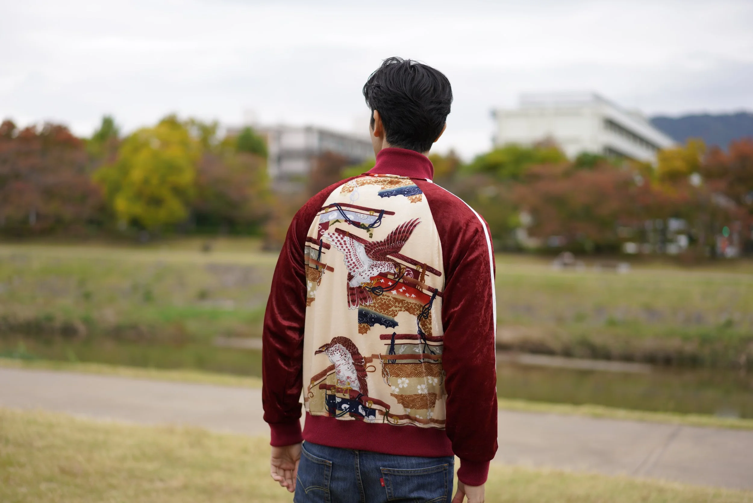 Back of a man standing outdoors near water, wearing a red and cream embroidered jacket with traditional Japanese motifs, with trees and buildings in the background.