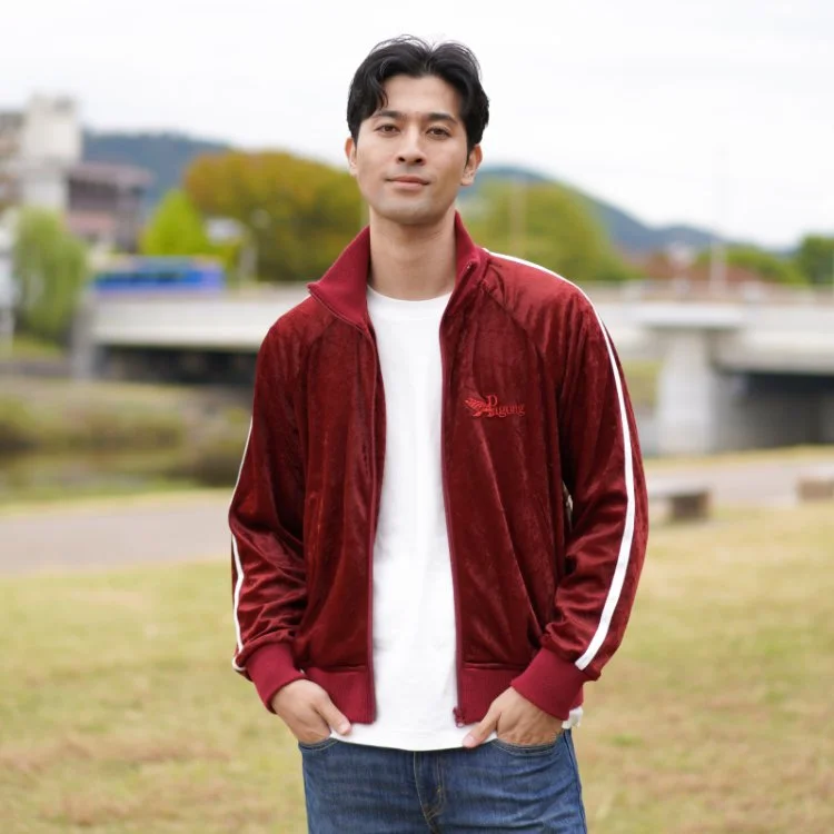 A young man wearing a red Velour Jacket and blue jeans, standing outdoors in a park with a river and bridge in the background.