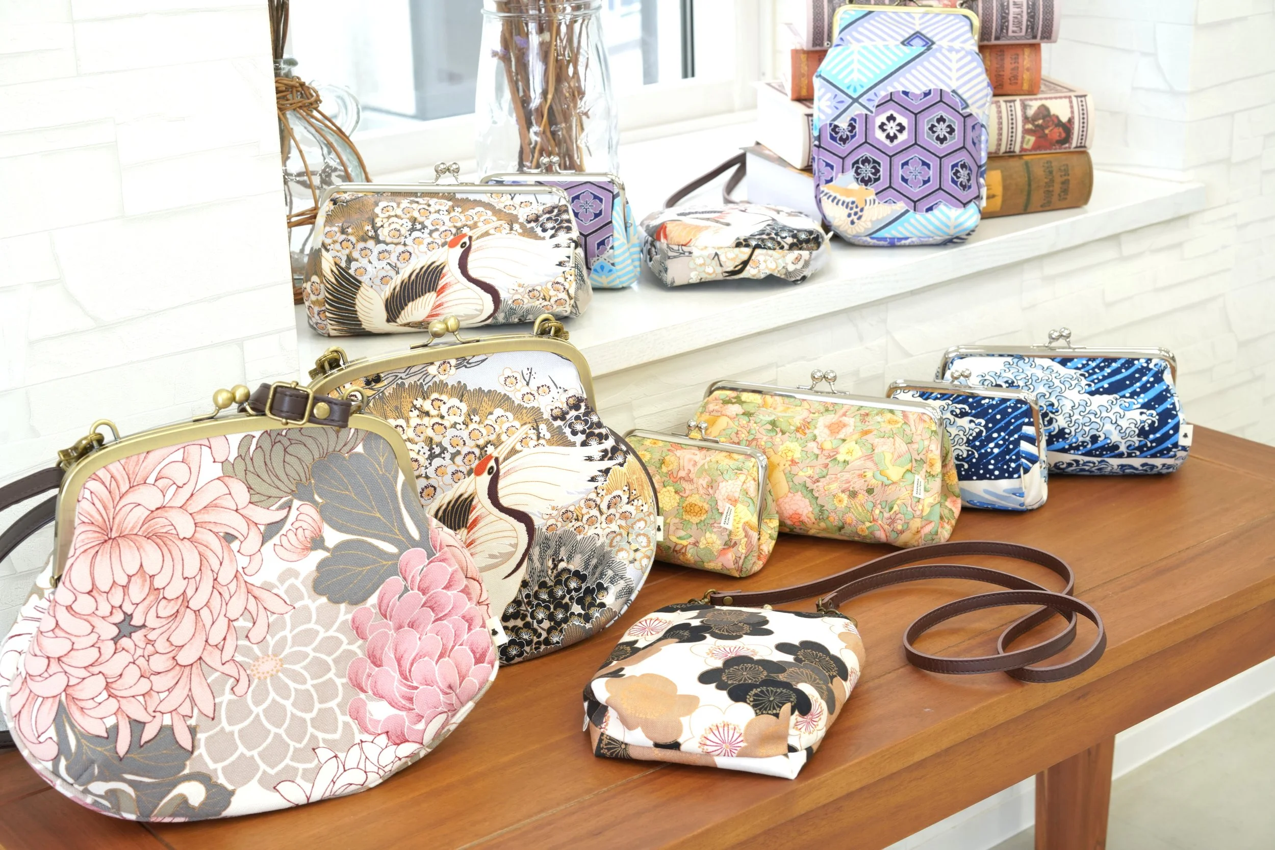 Display of various colorful floral and nature-themed handbags and pouches on a wooden table and shelf, with books in the background.