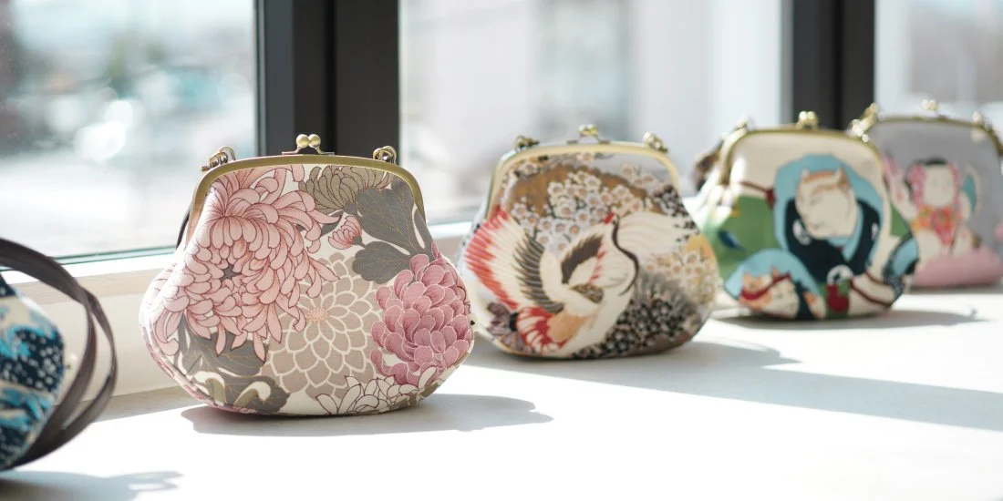 Hand made and hand dyed row of colorful patterned gamaguchi handbags displayed on a windowsill, with sunlight casting shadows.