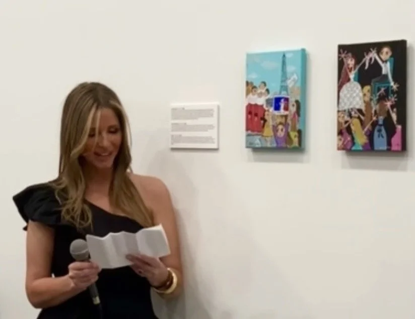 Artist Juliet Gilden speaking at an exhibition with her paintings displayed beside her.