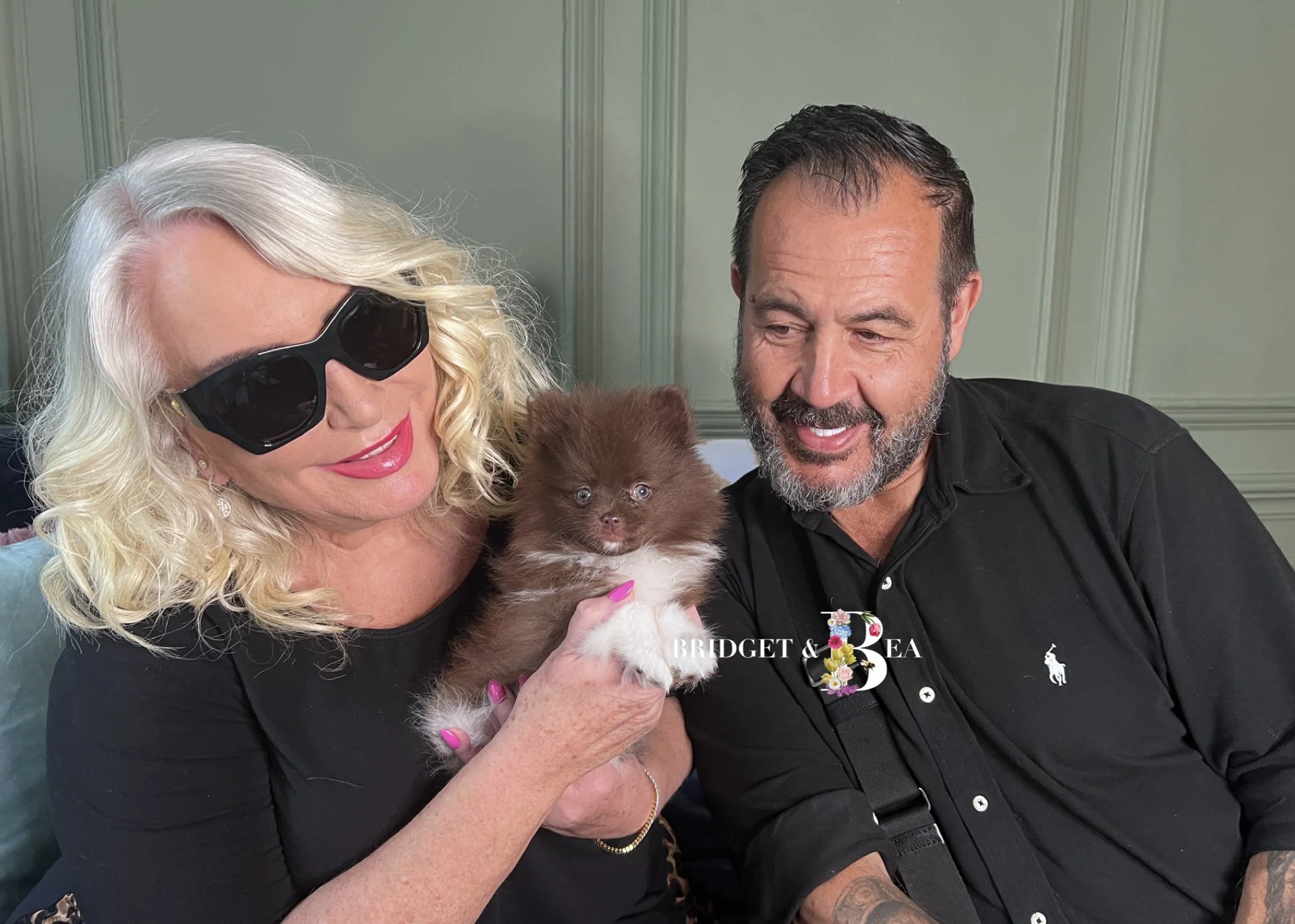 A woman with blonde hair, wearing black sunglasses and a black top, holds a small brown and white puppy. A man with dark hair and a beard, wearing a black shirt, sits beside her, all smiling and looking at the puppy. The background has green wall pan