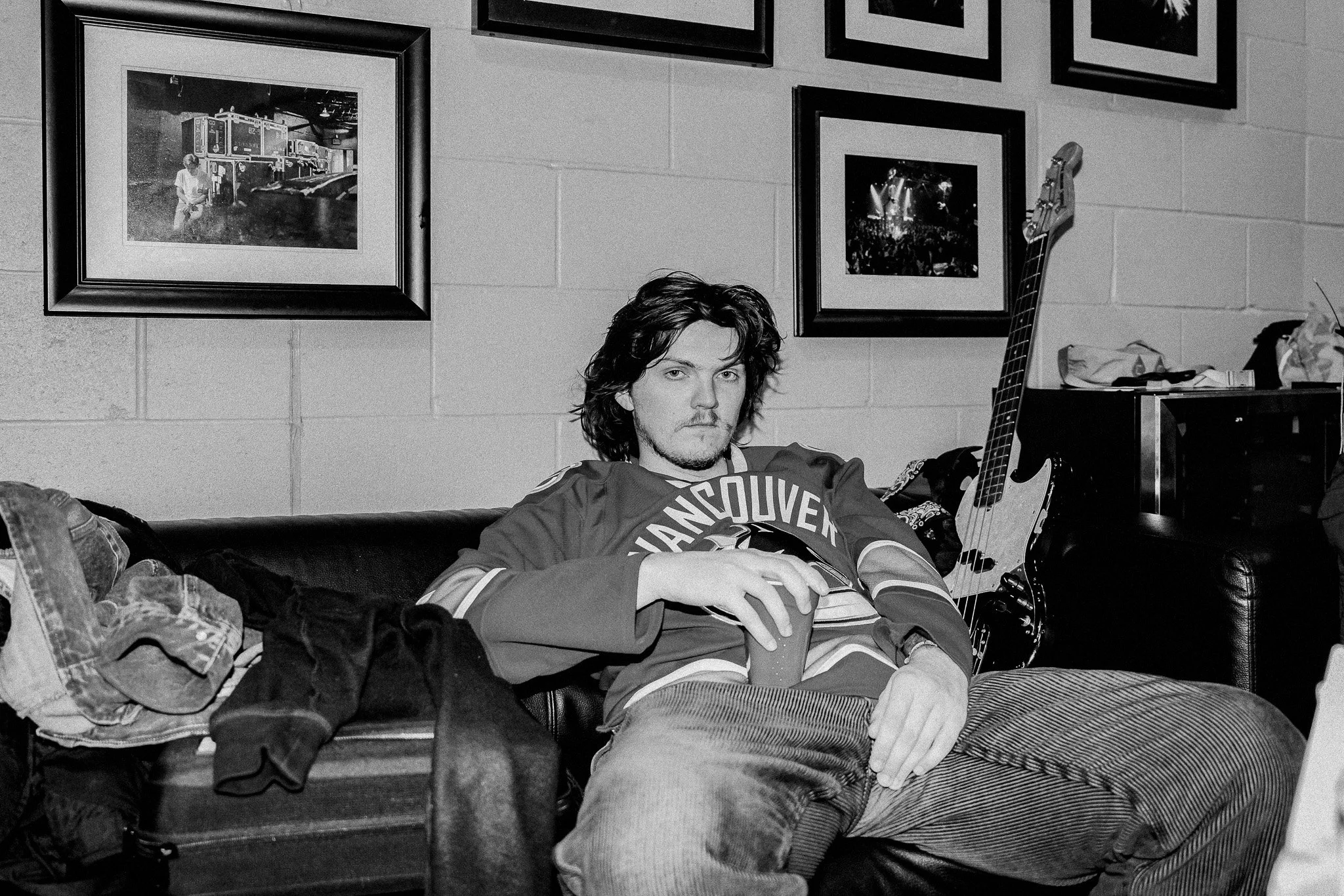 A young man with shoulder-length wavy hair sitting on a black leather couch in a room with framed photographs on the wall behind him. He is wearing a hockey jersey and holding a drink. An electric bass guitar is leaning against the wall to his right. There are clothes and a suitcase on the couch next to him.