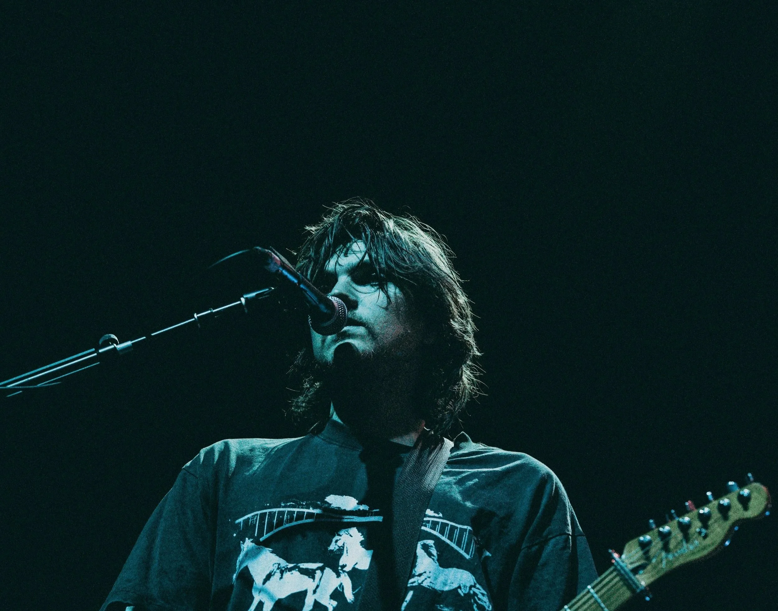 A male musician with long hair and a beard performs on stage, singing into a microphone and playing an electric guitar.