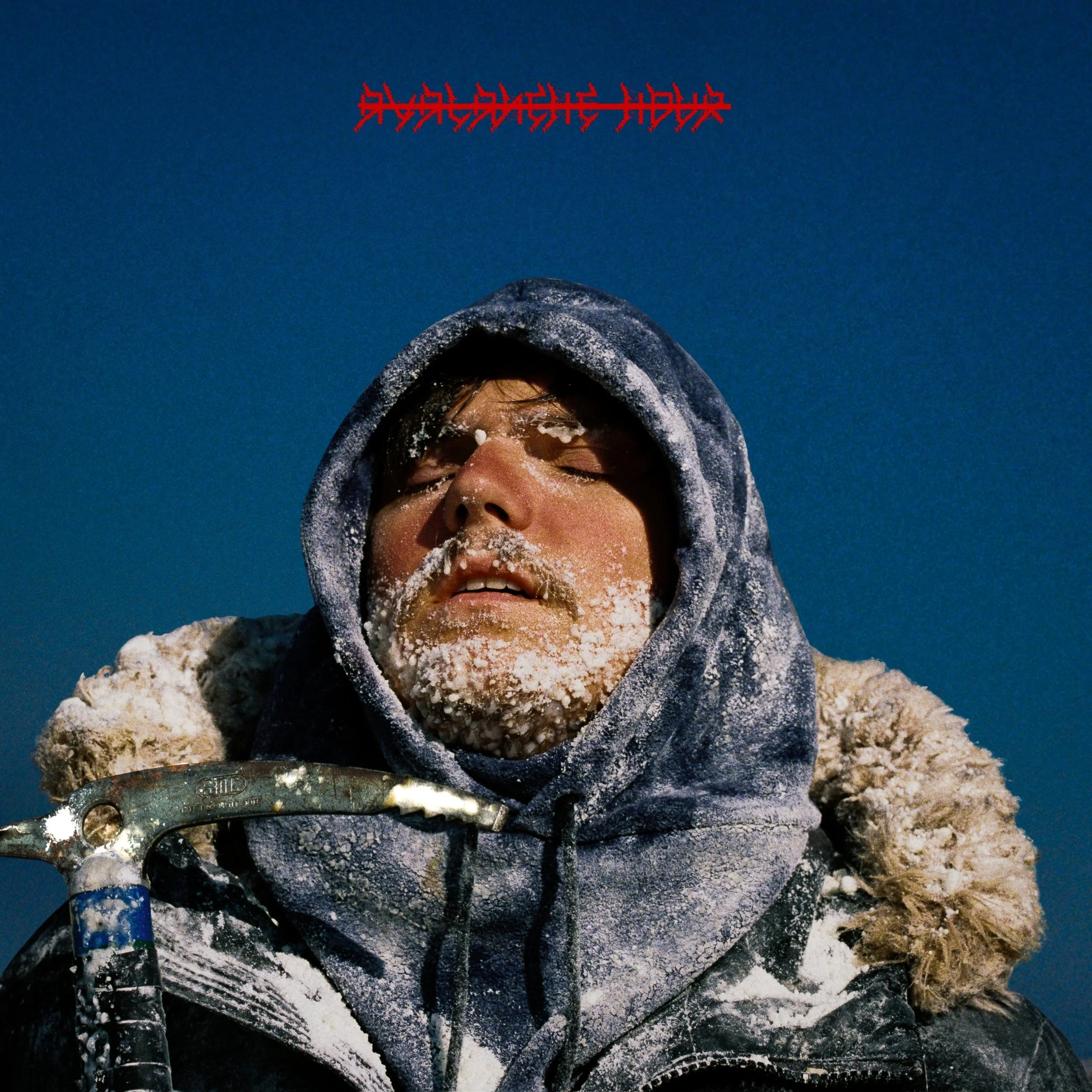 A person with frost and snow on their face and clothing, wearing a hooded jacket and holding an ice axe, in a cold, snowy environment.