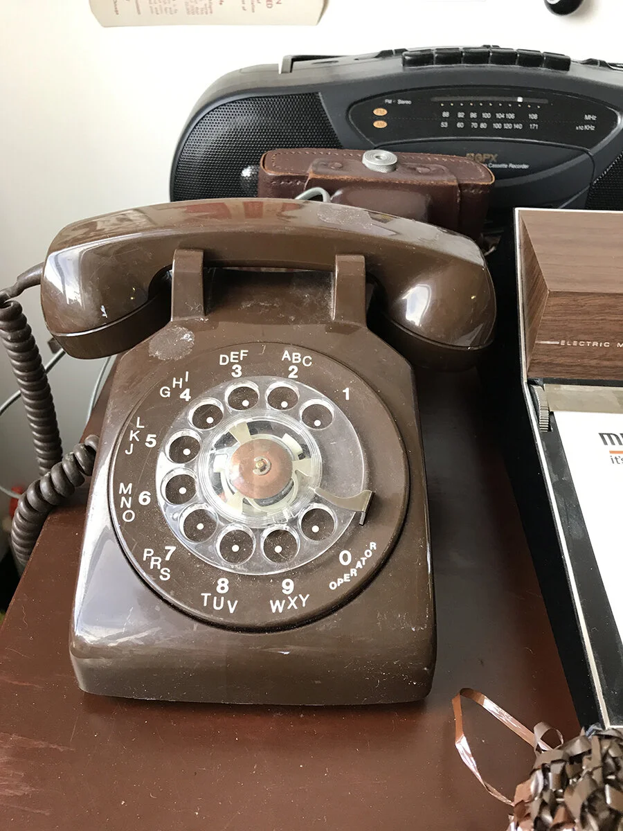 My plan for the exhibit was to carry the objects I had created in the pockets of the trench coat, as a sort of “mobile” exhibition. In place of an actual exhibition, this telephone on the desk would sit in the hallway gallery. A hidden camera would …