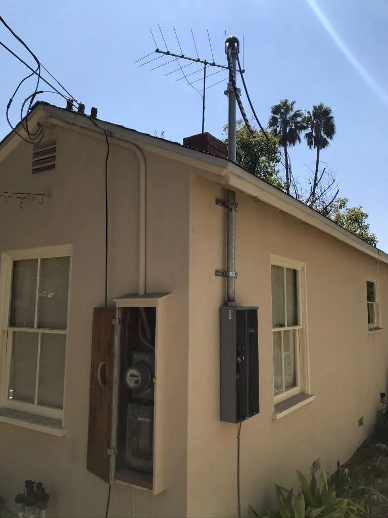 Side view of a beige house with electrical meters and a grey electrical panel outside, with an antenna on the roof and palm trees in the background.