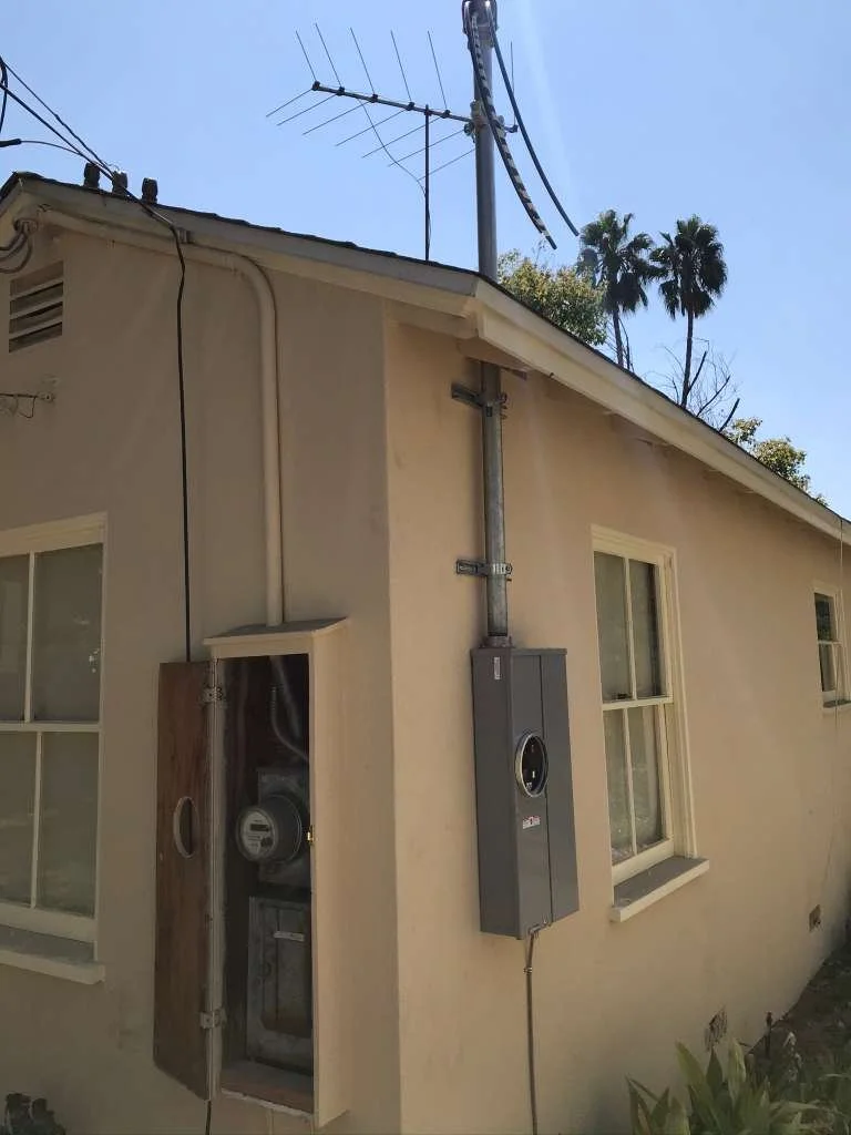 Exterior of a house with an open electrical meter box, electrical panel, and various wires and antenna on the roof. Two windows are visible on the side of the house.