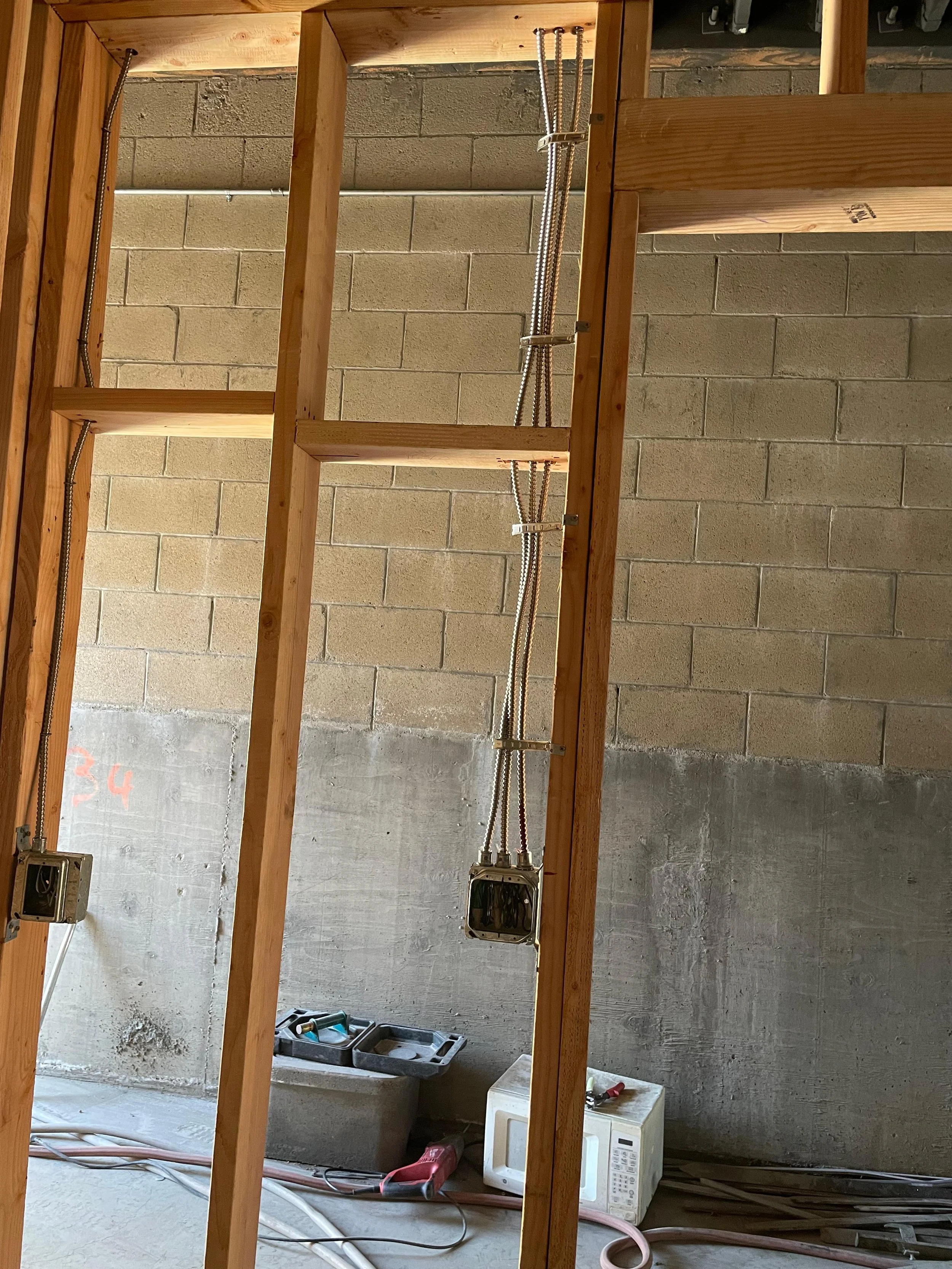 Construction site with wooden framing and electrical wiring, unfinished concrete wall, electrical boxes, and plumbing pipes on the floor.