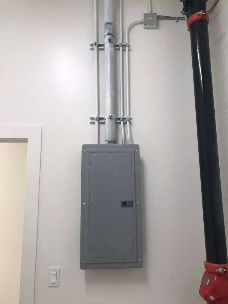 Electrical and plumbing conduit pipes installed on a wall with an electrical panel and a large black pipe.