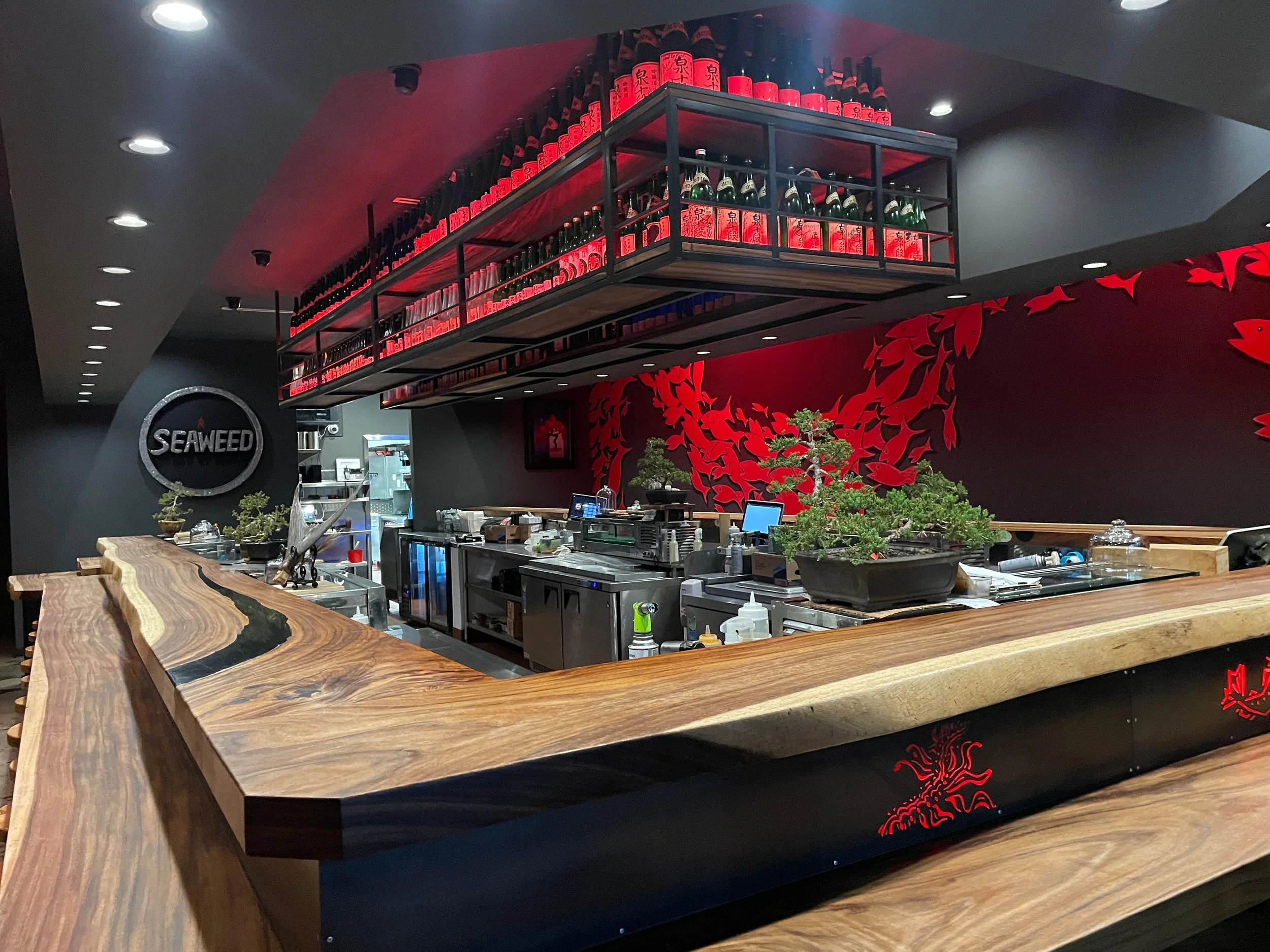 A Japanese-themed restaurant counter with a woodgrain countertop and a black wall featuring red decorative fish and dragons. Shelves above hold bottles of sake and wine, with a small bonsai tree on the counter.