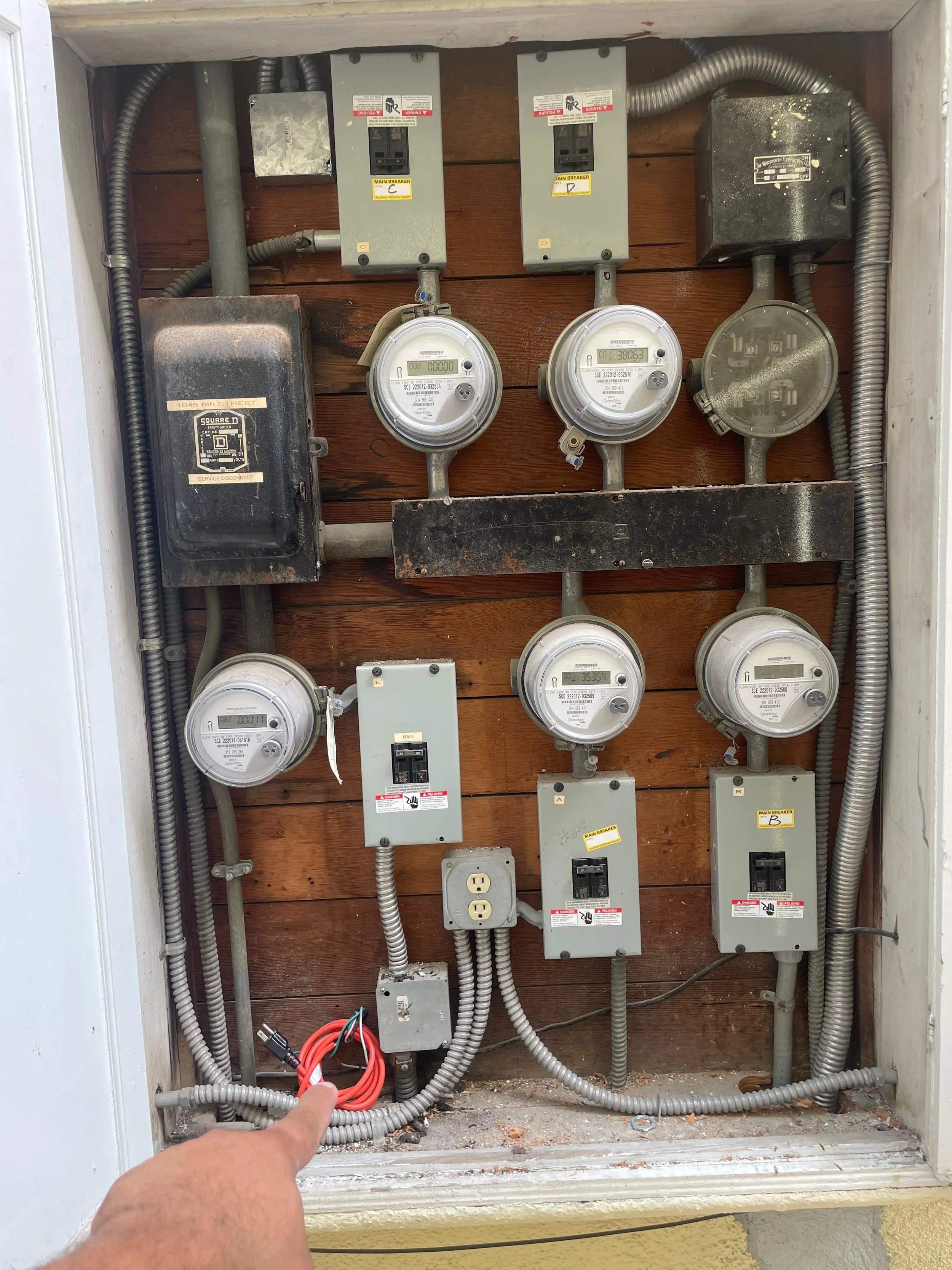 An open electrical panel with multiple electrical meters, circuit breakers, and wiring. A person's hand is holding a bundle of red and black electrical wires at the bottom of the panel.